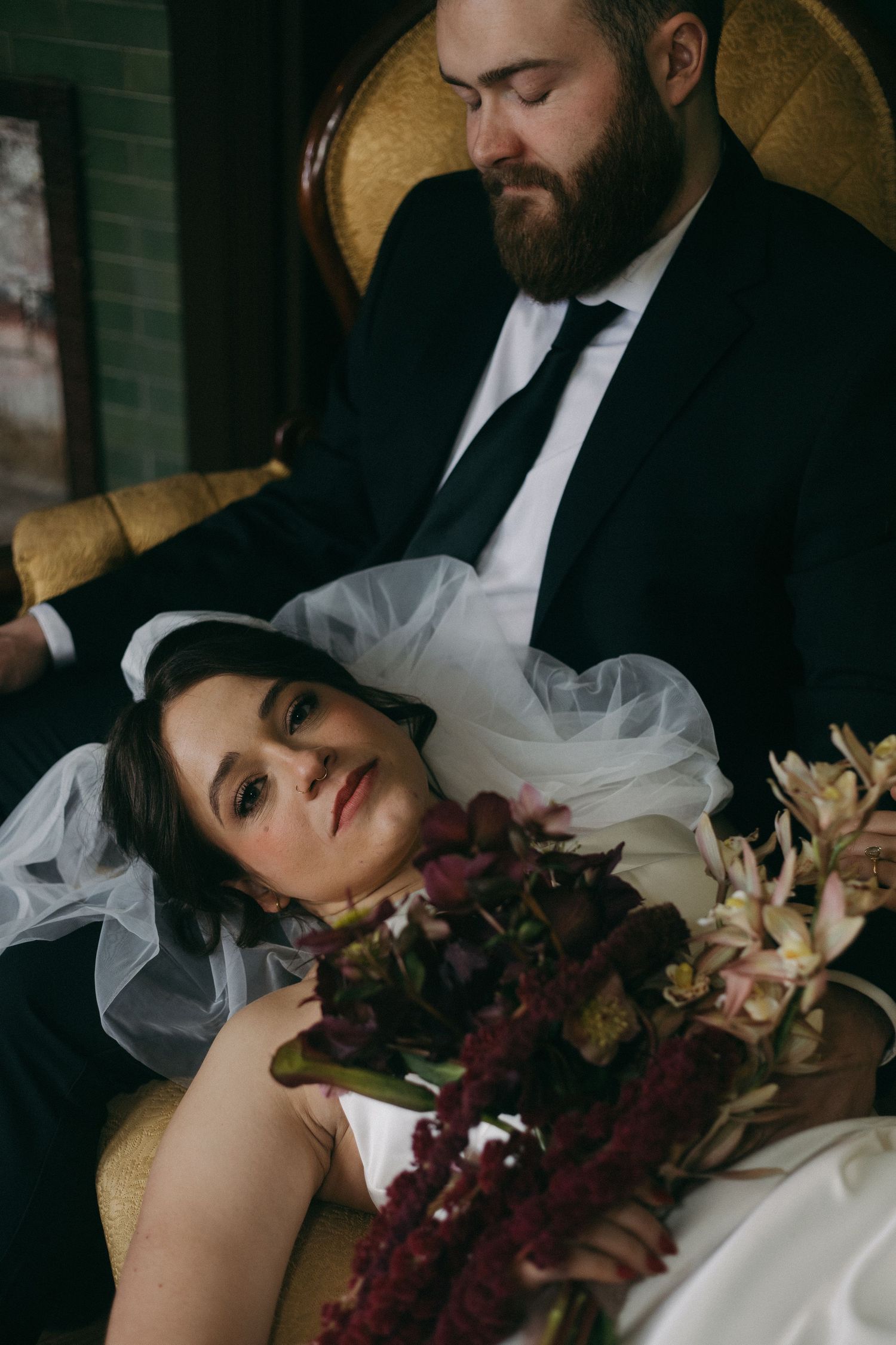 A couple in wedding attire share an intimate moment in a vintage chair with a dark burgundy bouquet.