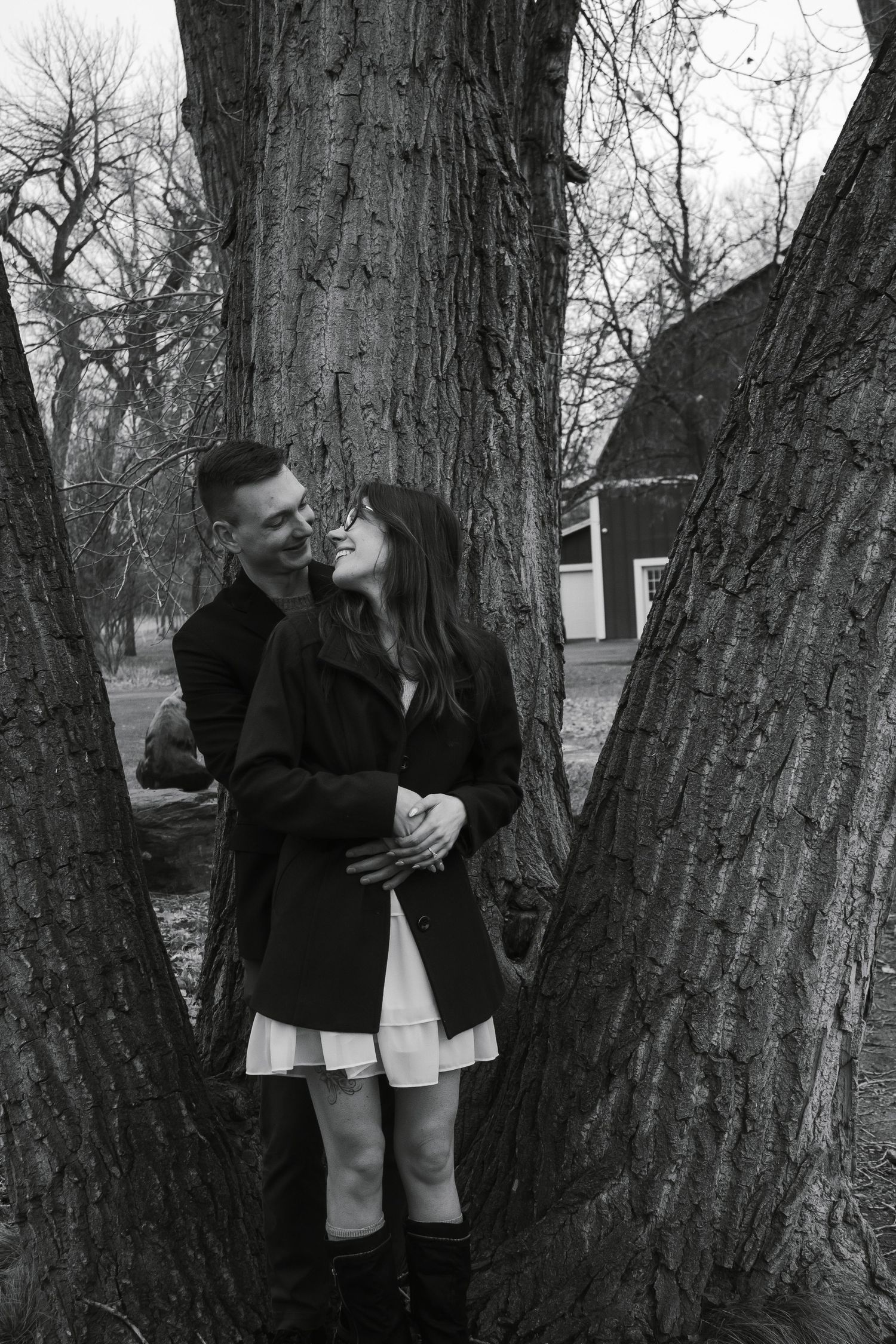 A couple sharing an intimate moment between two large tree trunks in black and white.