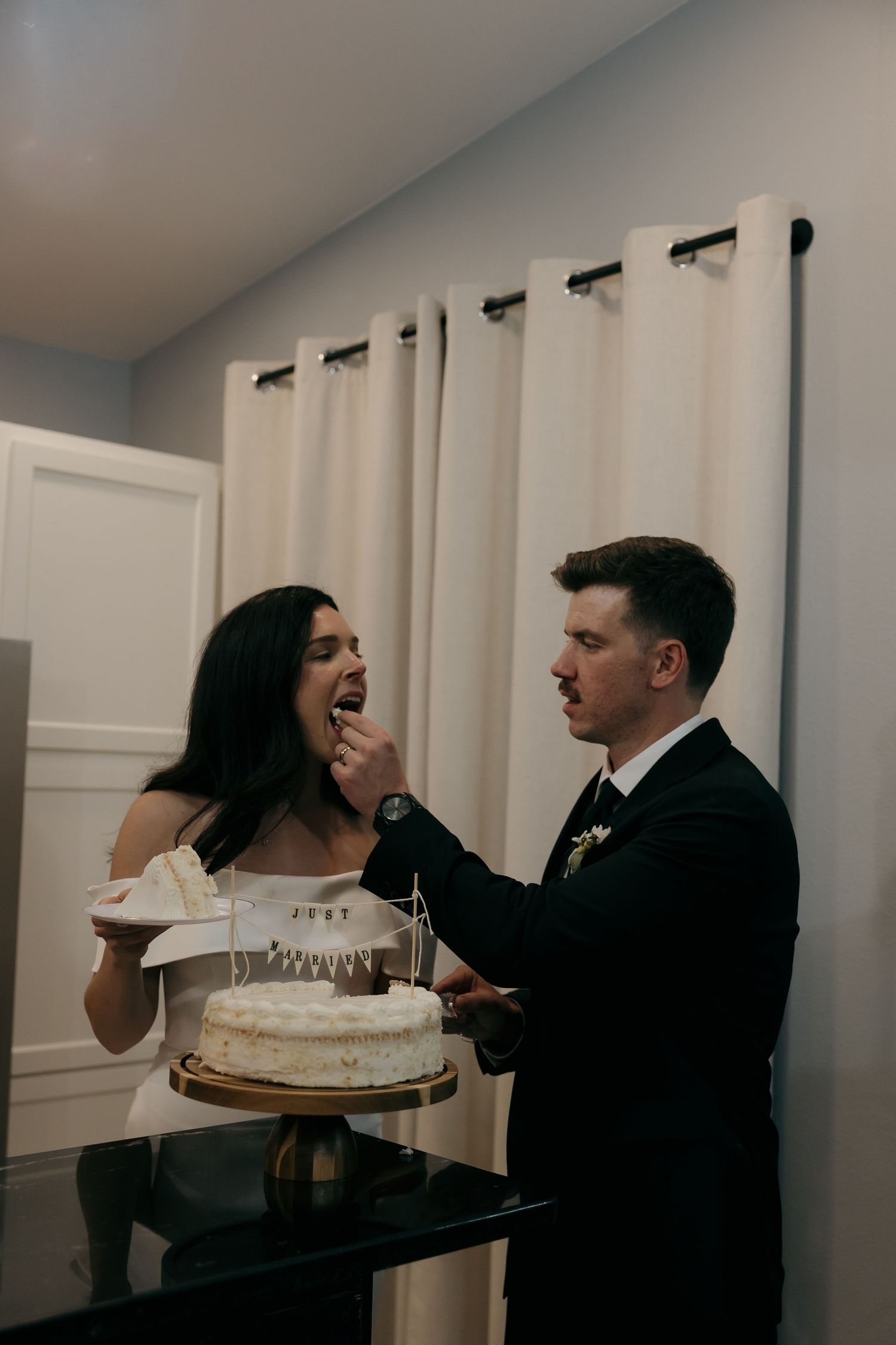 Groom feeds wedding cake to his smiling bride during their intimate indoor cake cutting ceremony.