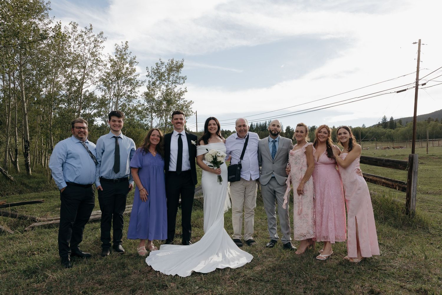 Wedding party poses outdoors on green lawn with trees, bride in white gown surrounded by family in formal attire.