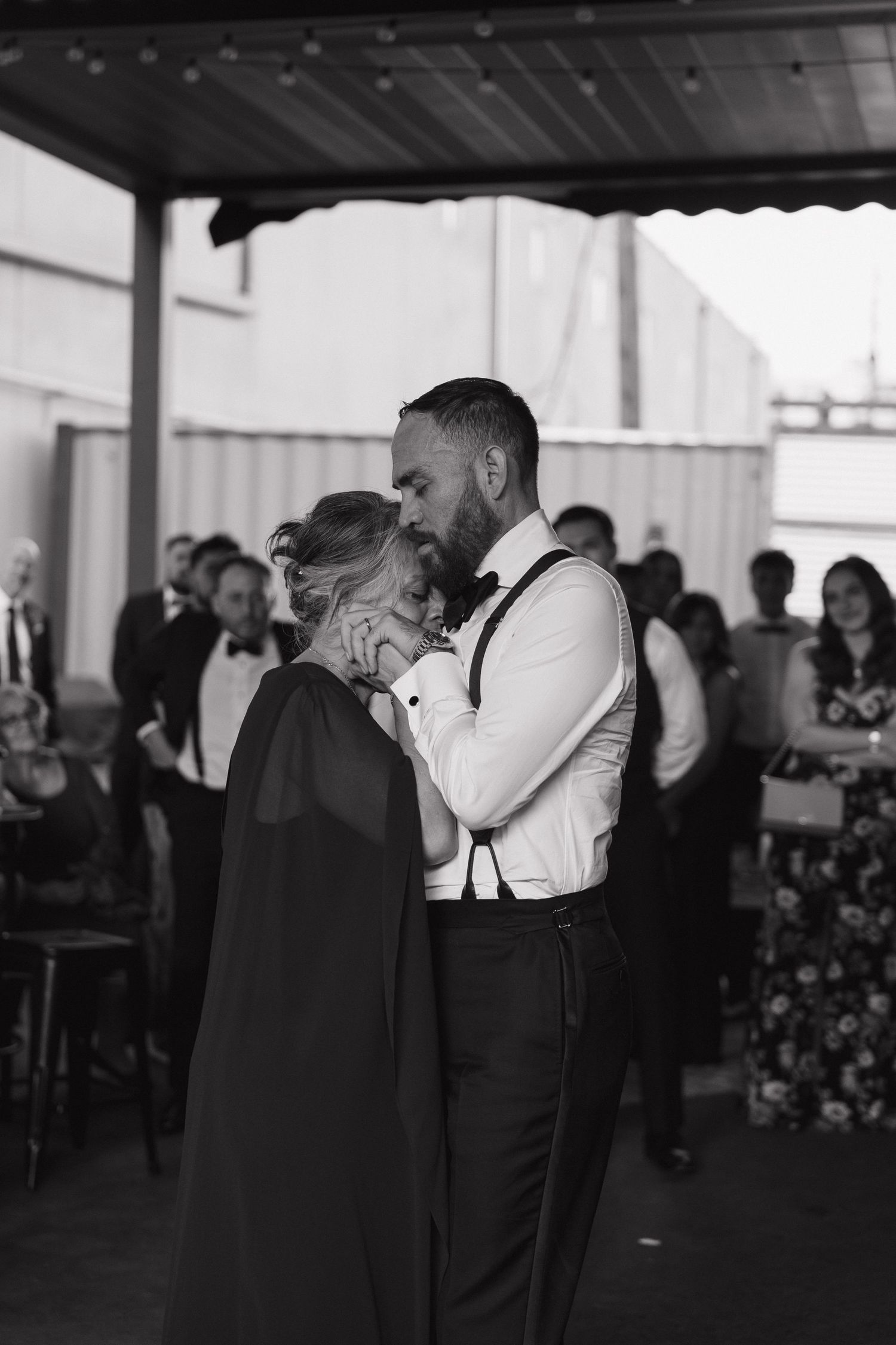 Black and white photo of groom dancing with woman in cape at wedding reception with guests.