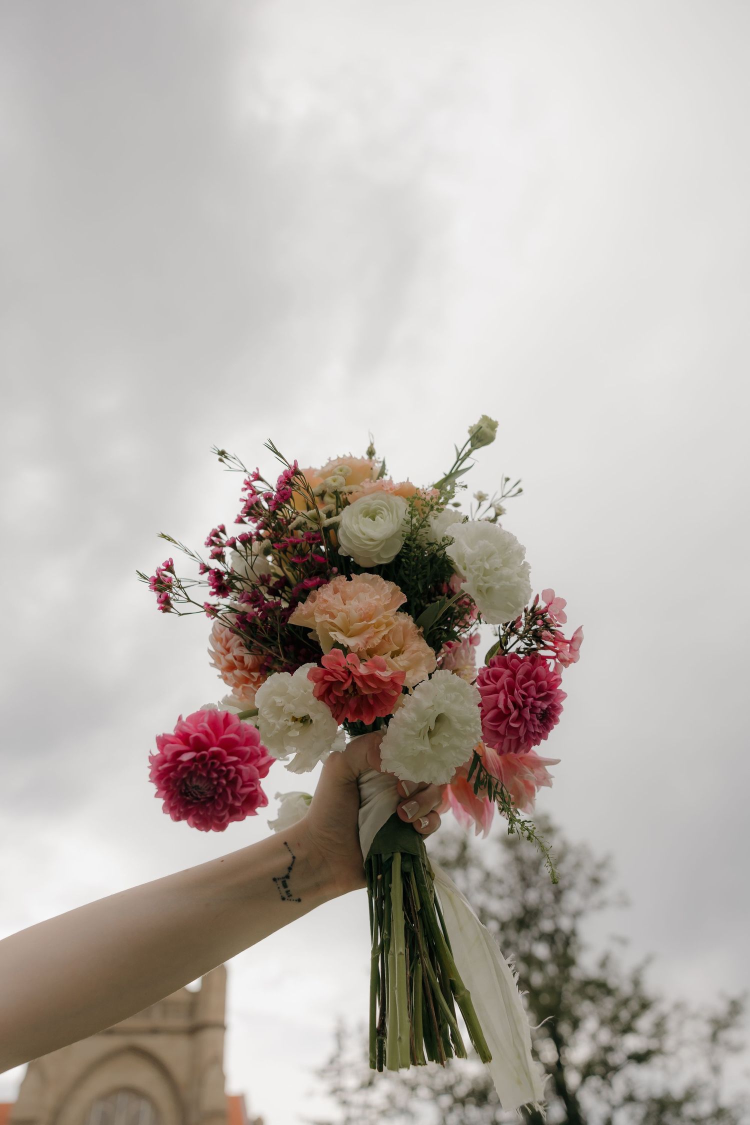 Bride holding a colorful wedding bouquet of pink, coral, and white flowers raised against a cloudy sky.