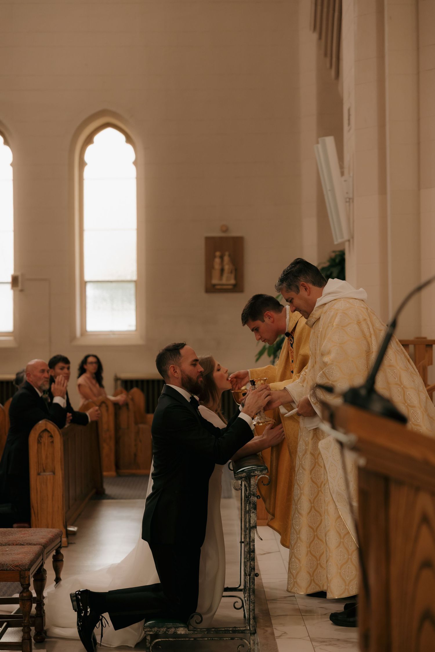Bride and groom kneel at altar rail receiving blessing from priest during Catholic wedding Mass.