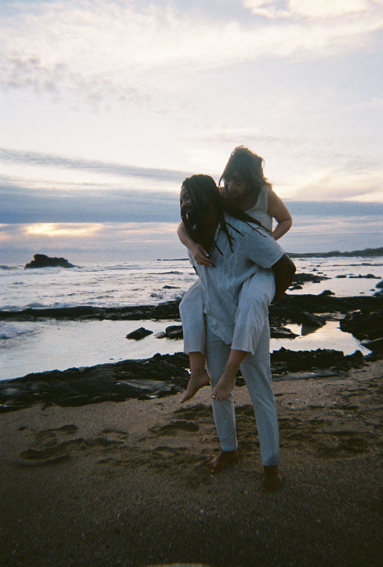 A couple in white clothing embraces on a rocky beach at sunset with ocean waves and cloudy skies in the background.