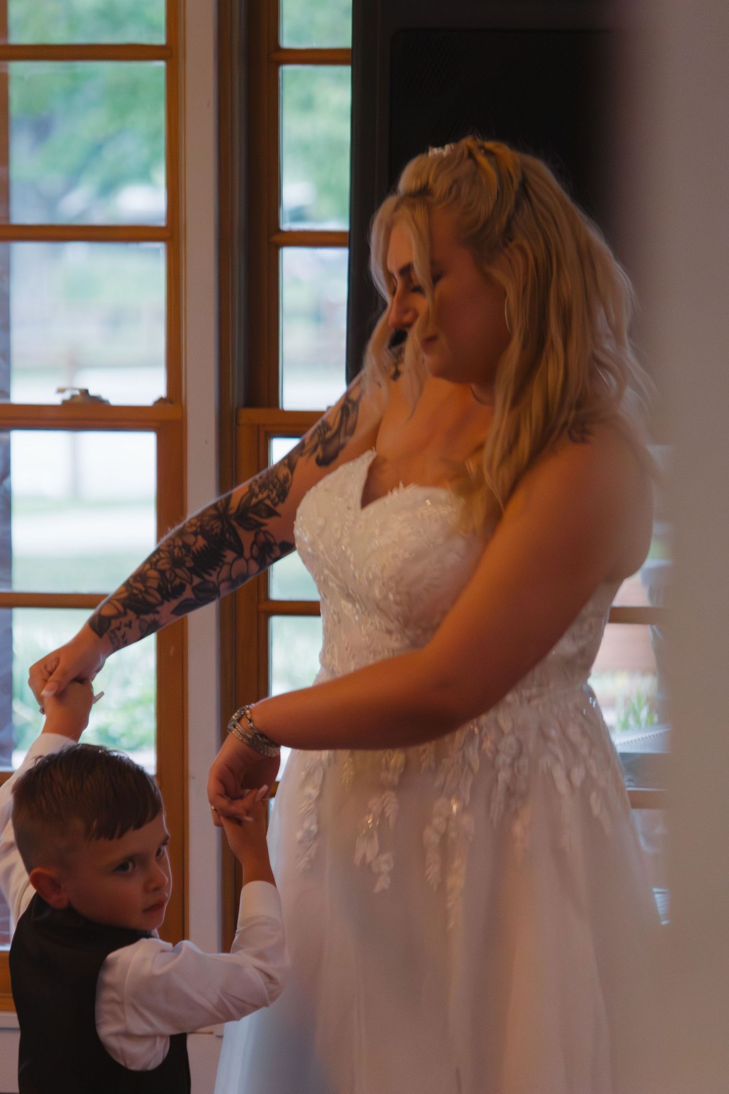 A bride in a white gown shares a tender dance moment with a young boy by window light.