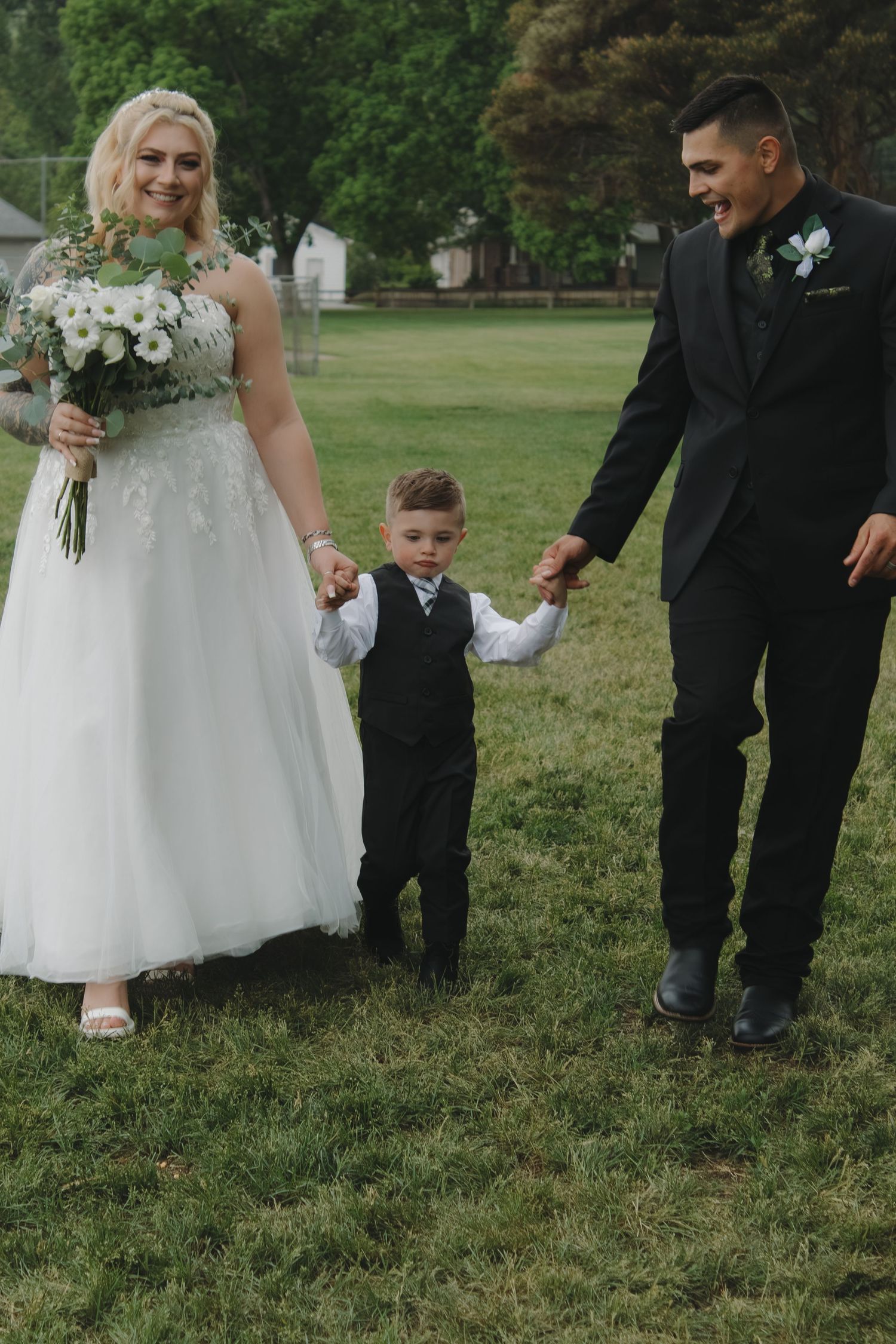 Wedding couple shares playful moment with young ring bearer on green lawn.