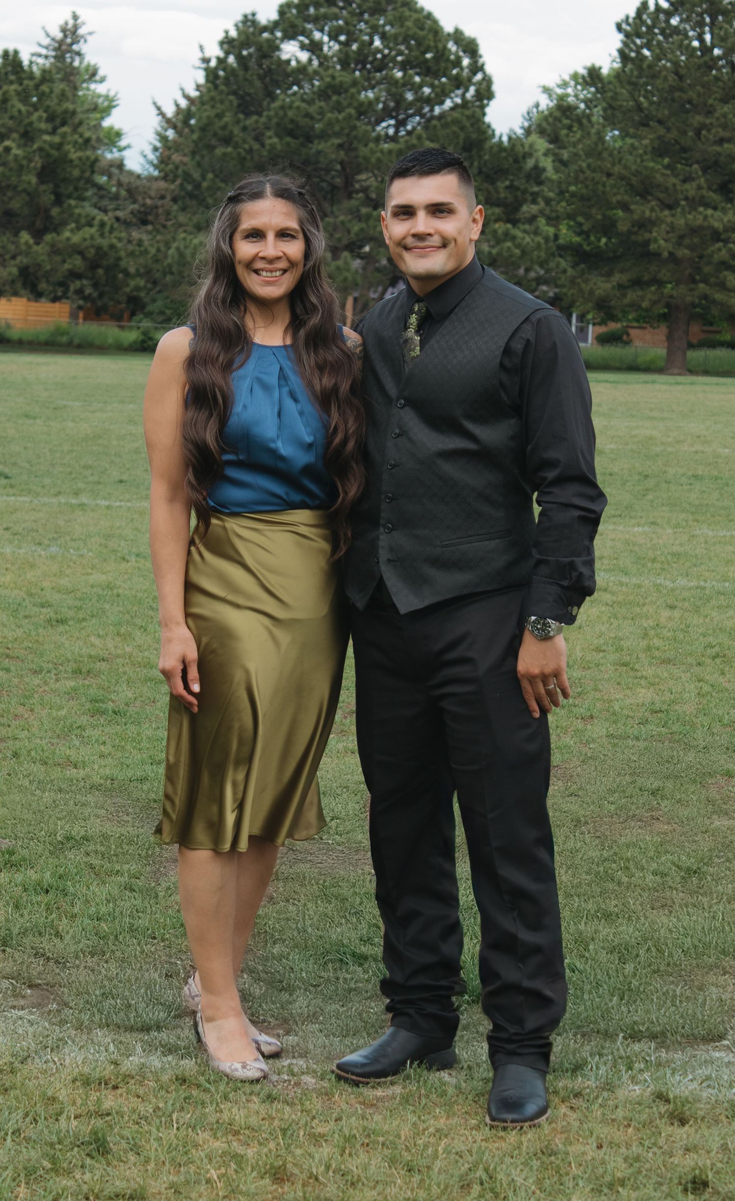 A couple poses together outdoors, with one wearing a blue top and gold skirt.