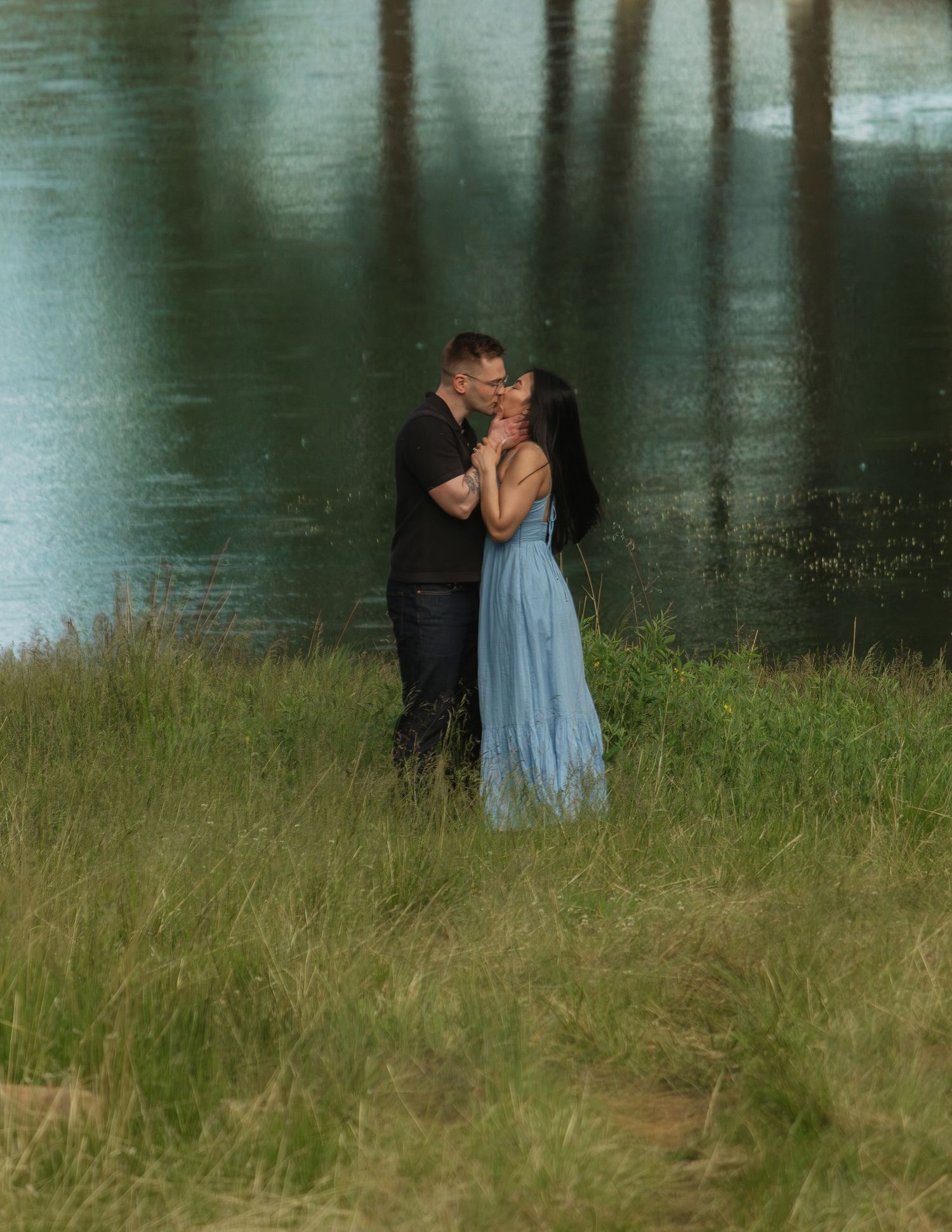 A romantic moment captured by a lake with tall grass in the foreground and reflective water in the background.