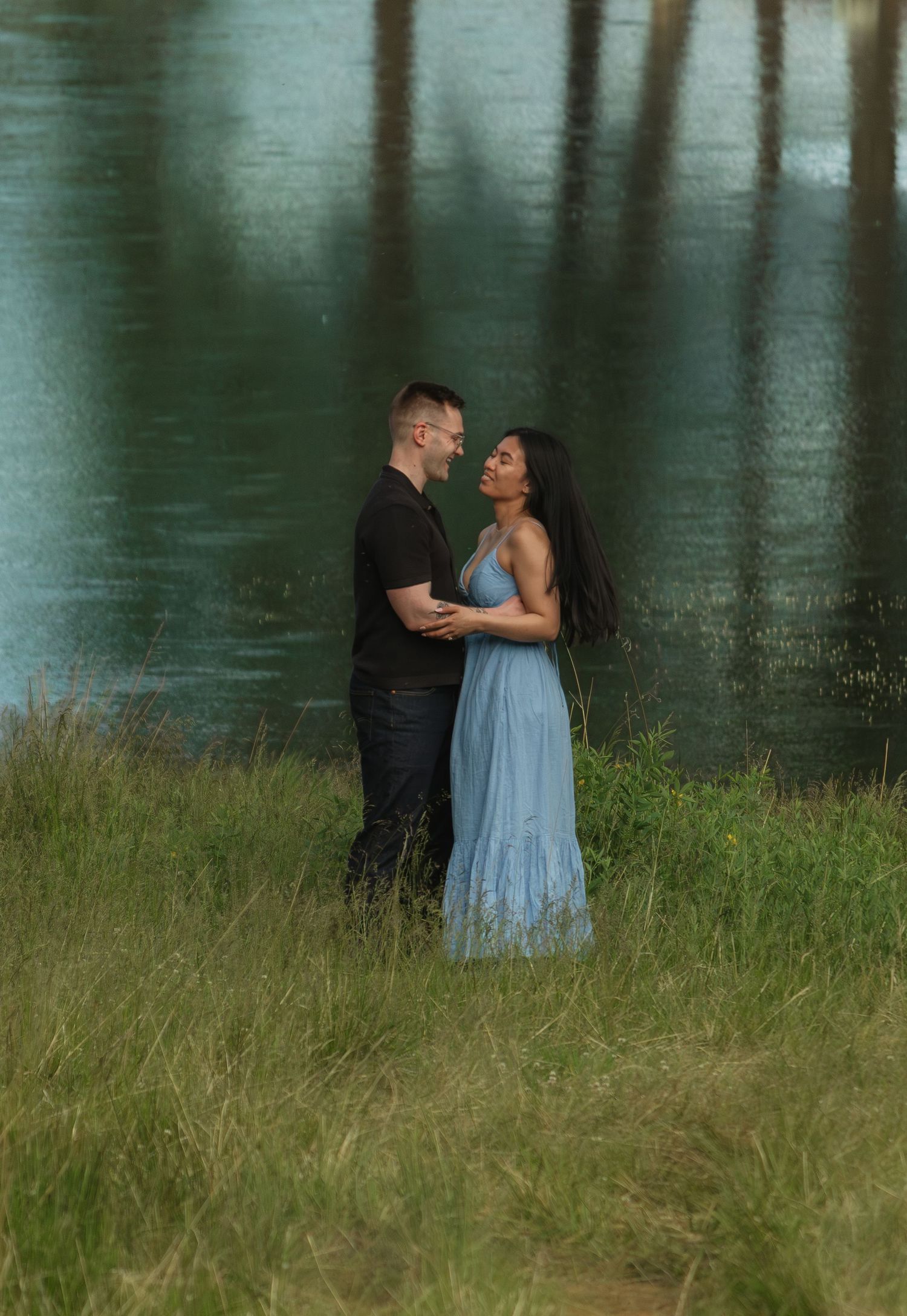 Romantic lakeside scene with two people in casual attire embracing in tall grass.