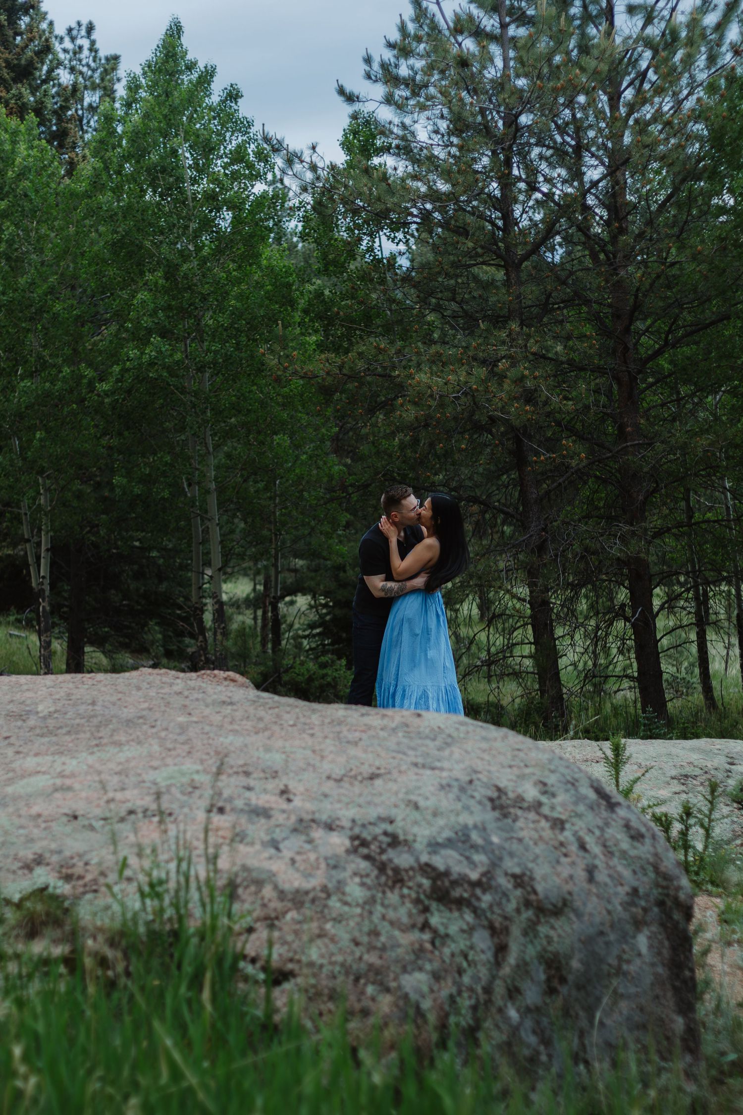 A couple embraces on a large boulder surrounded by pine trees at dusk.