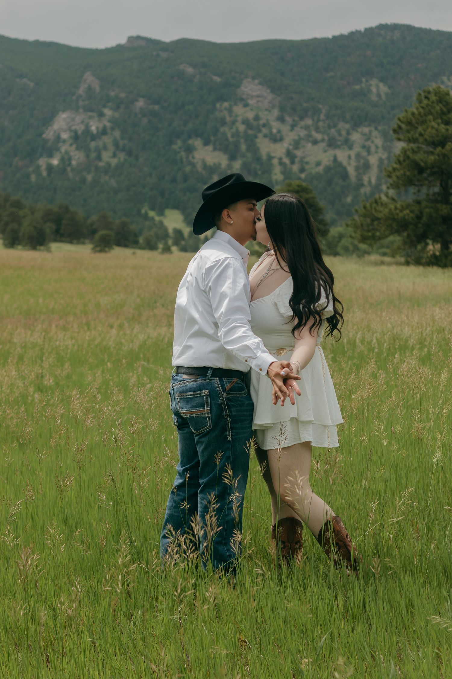 Romantic couple in western attire standing close in a mountain meadow with tall grass and pine trees.