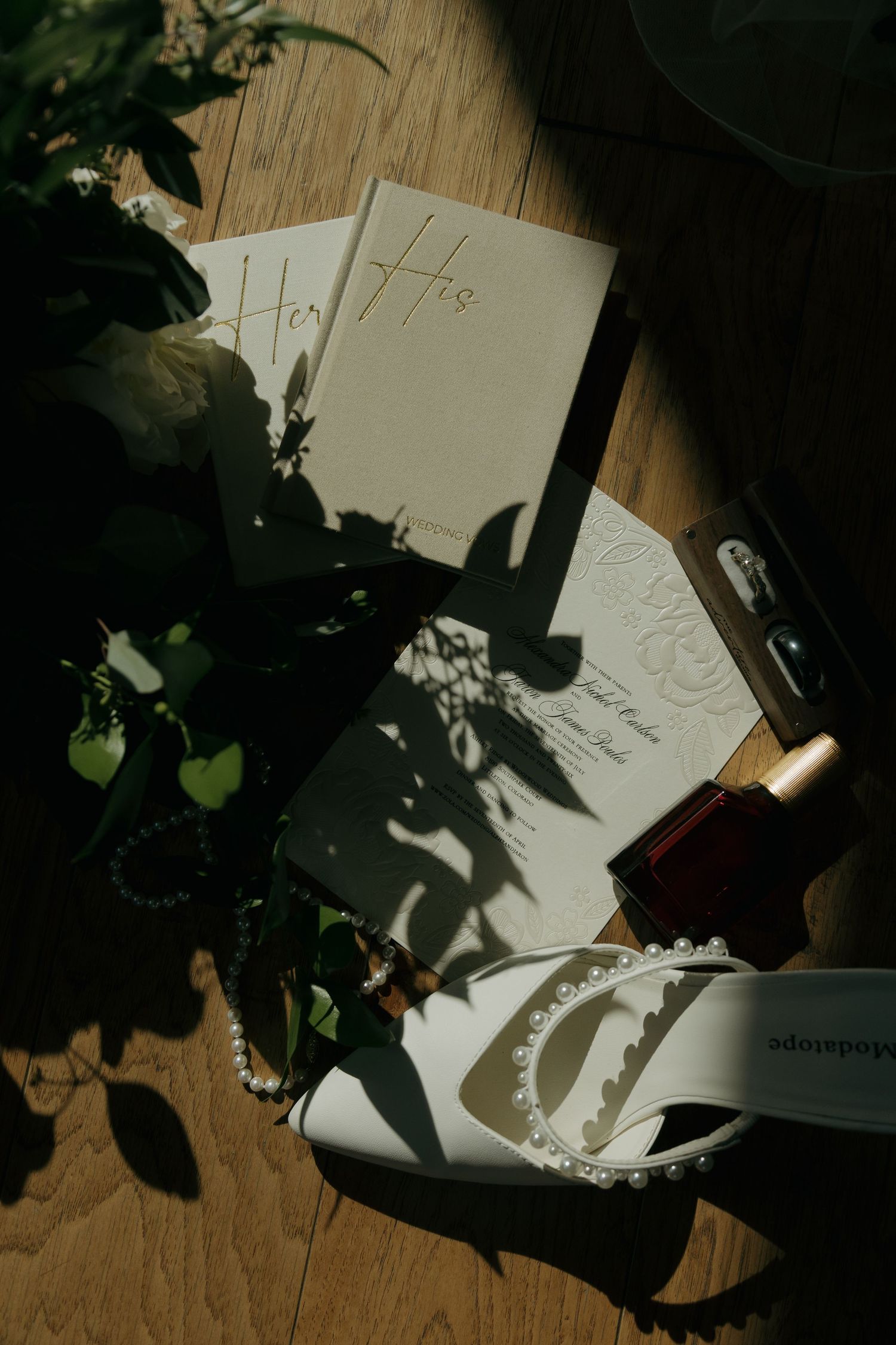 Wedding flat lay with vow books, bridal heels, pearl jewelry, and florals in dramatic light.