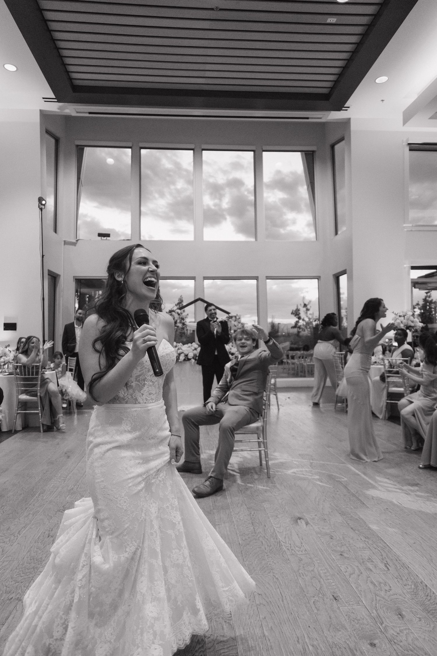 Bride in mermaid gown laughs while singing into microphone during black and white wedding reception.