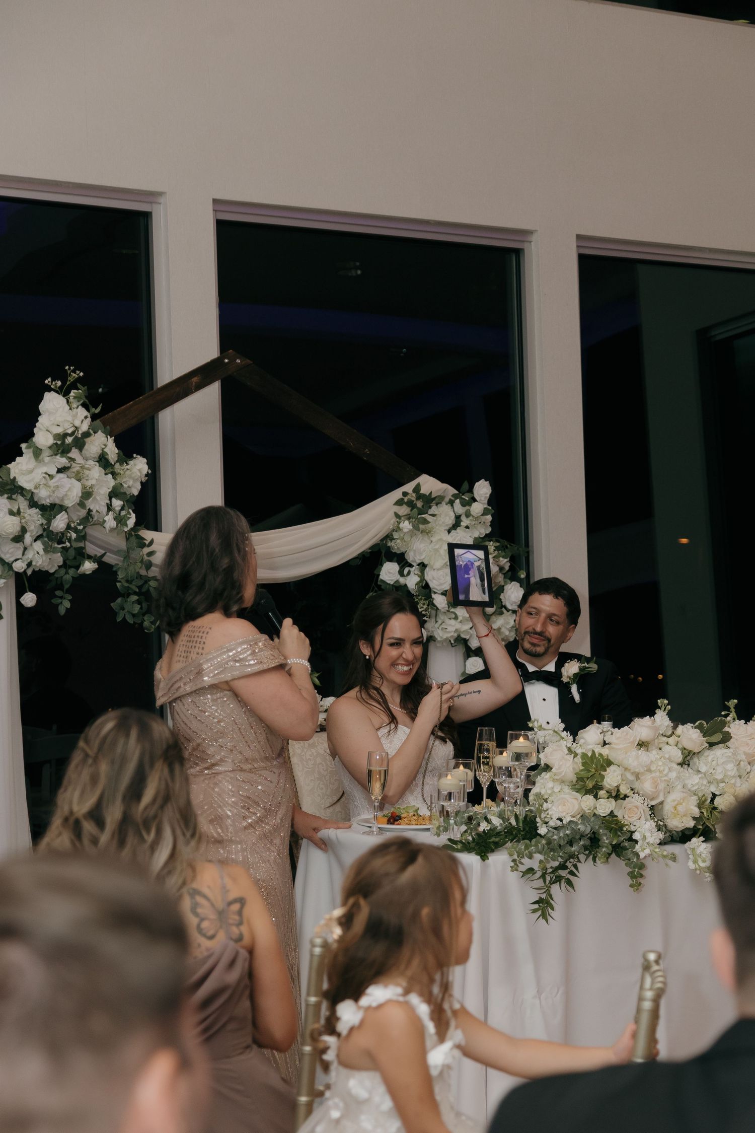 Bride and groom smiling at sweetheart table surrounded by white floral arrangements during wedding reception.