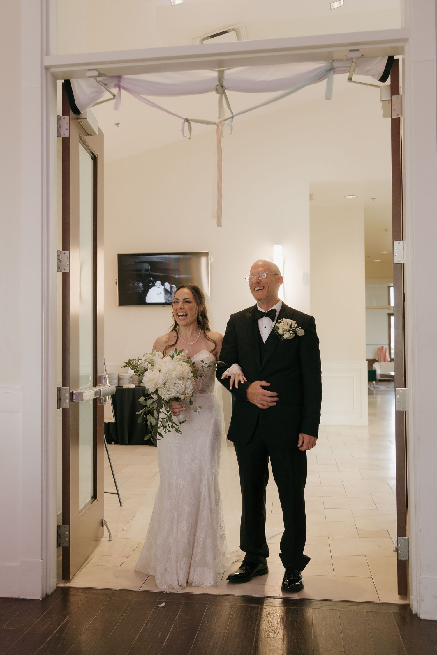 Smiling bride in lace gown walks with father in tuxedo through elegant wedding venue entrance.