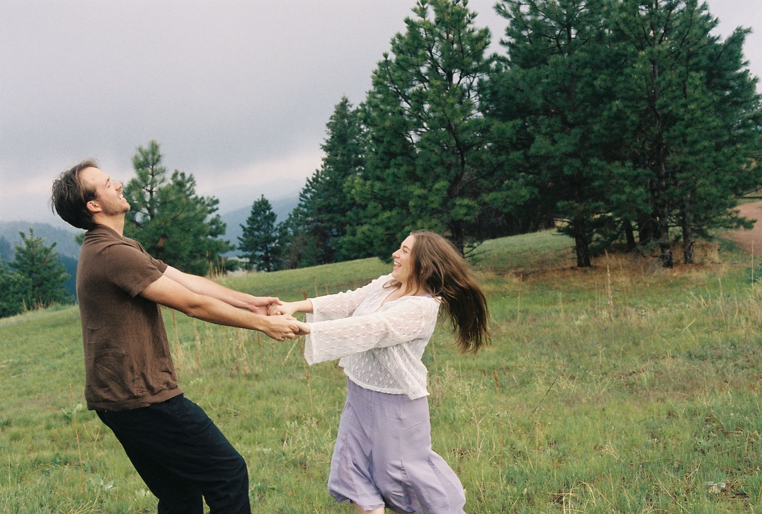 Couple spinning and laughing together in a green meadow with pine trees under an overcast sky.