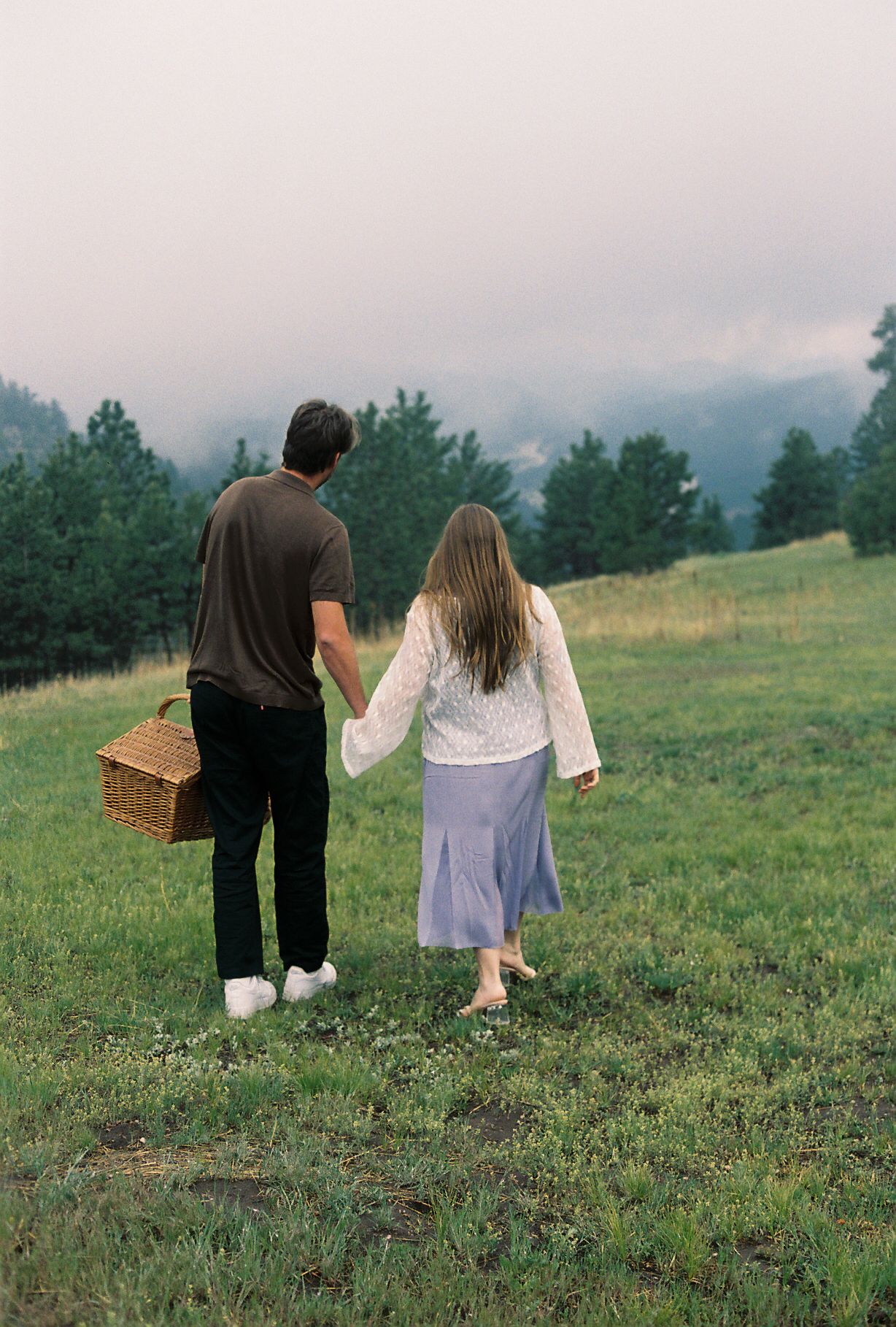 Couple holding hands walking through green meadow with picnic basket, misty mountains and pine trees in background.