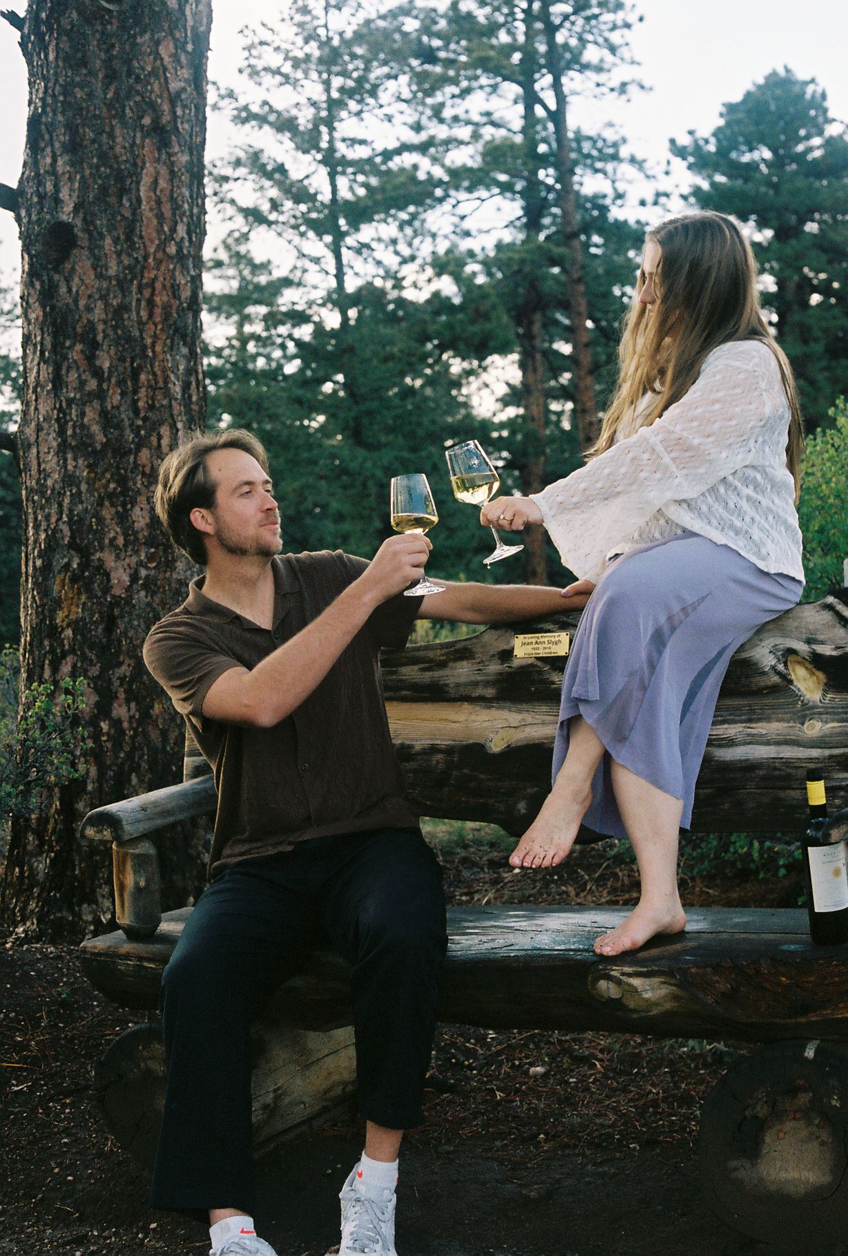 Couple toasting with white wine glasses on a rustic log bench surrounded by tall pine trees.