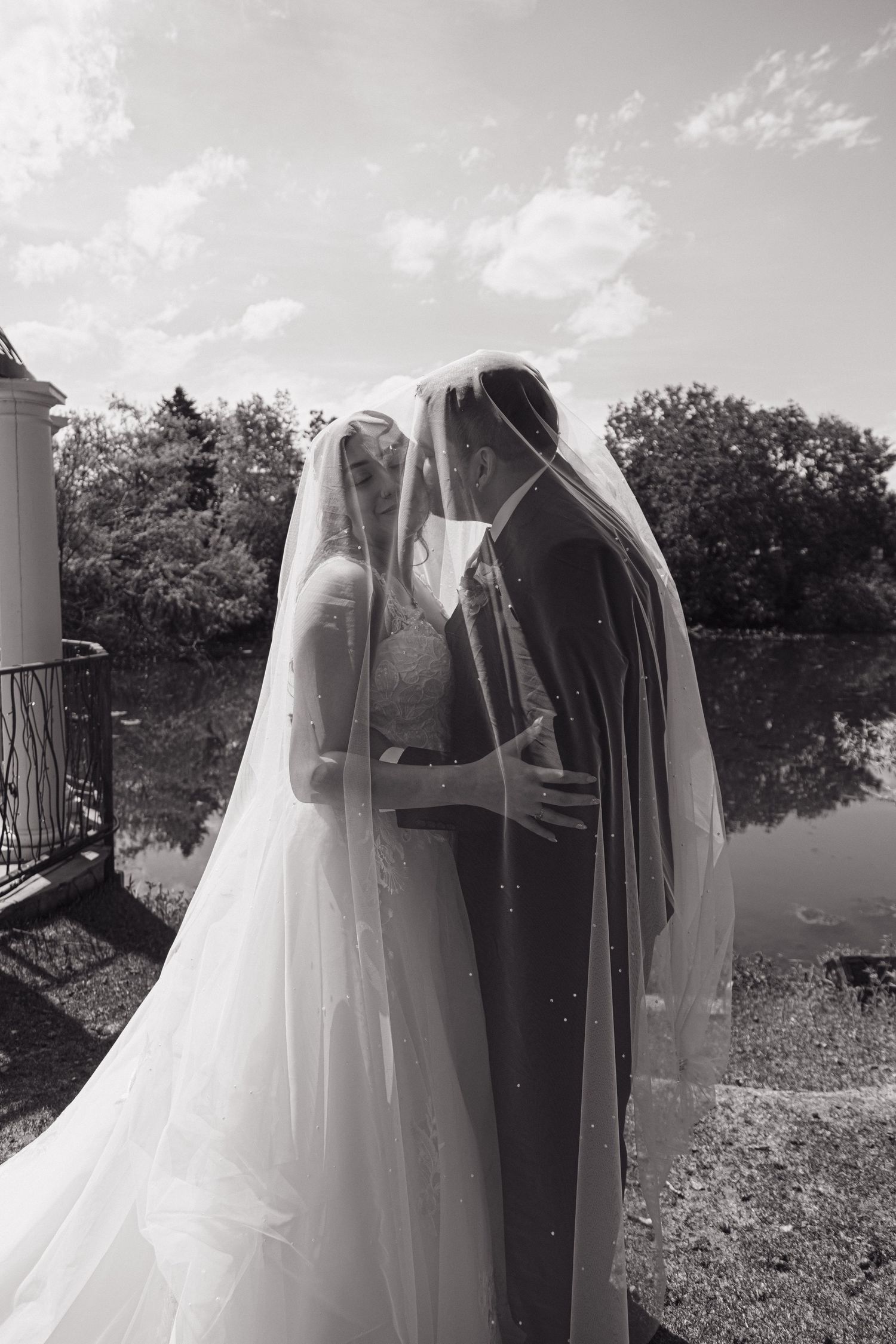 Black and white romantic photo of bride and groom sharing a kiss under a flowing wedding veil outdoors.