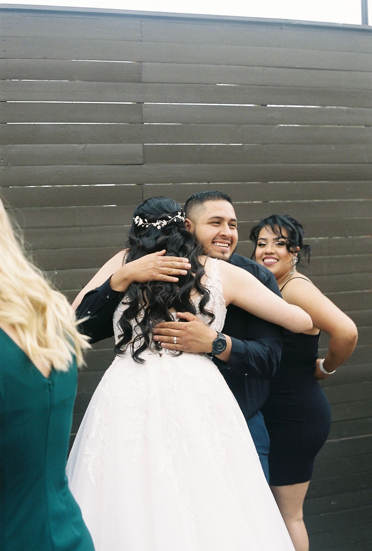Groom joyfully embraces bride and smiling guests outside during wedding celebration on film photo.