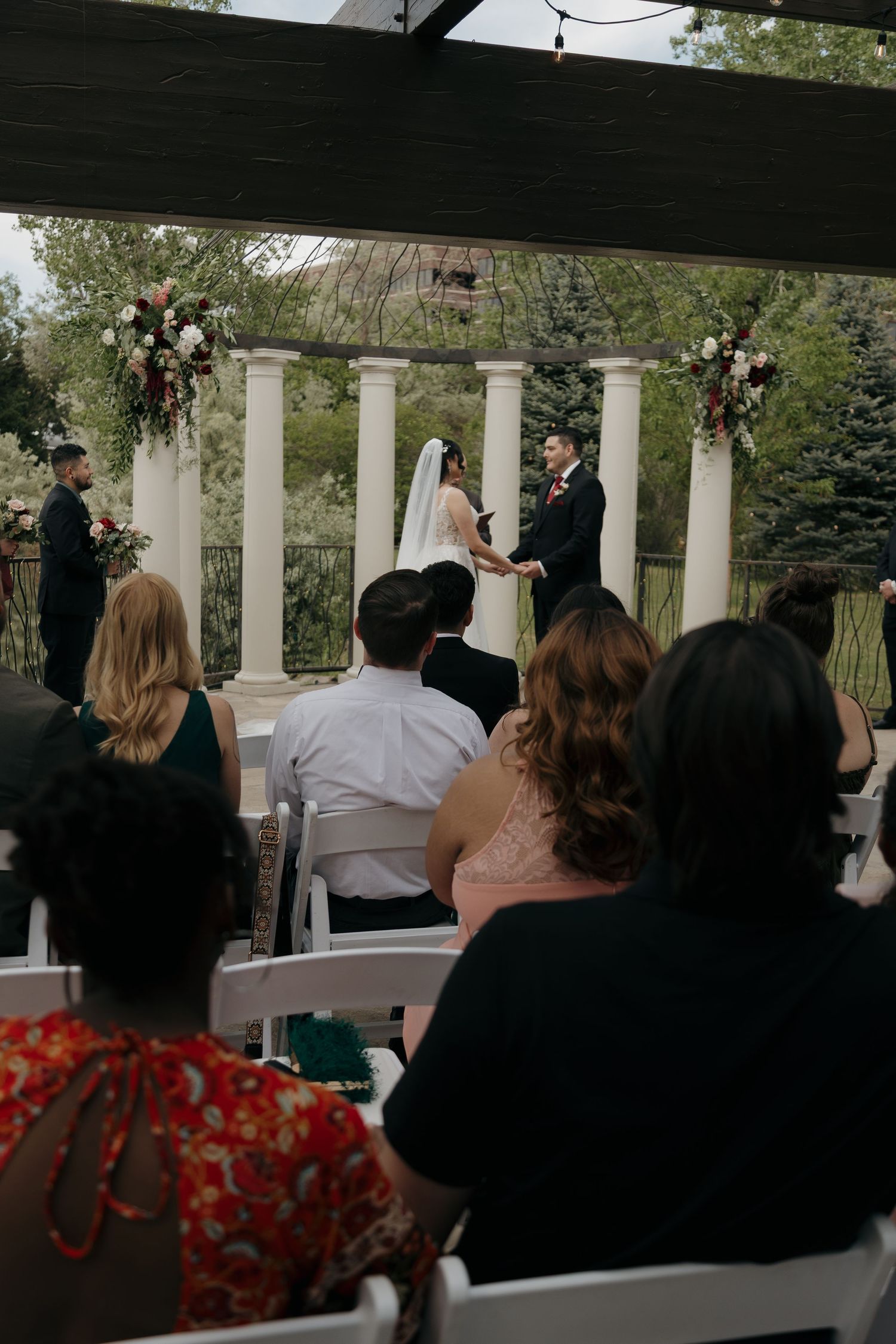 Bride and groom exchange vows at outdoor wedding ceremony beneath pergola with white columns and floral arrangements.