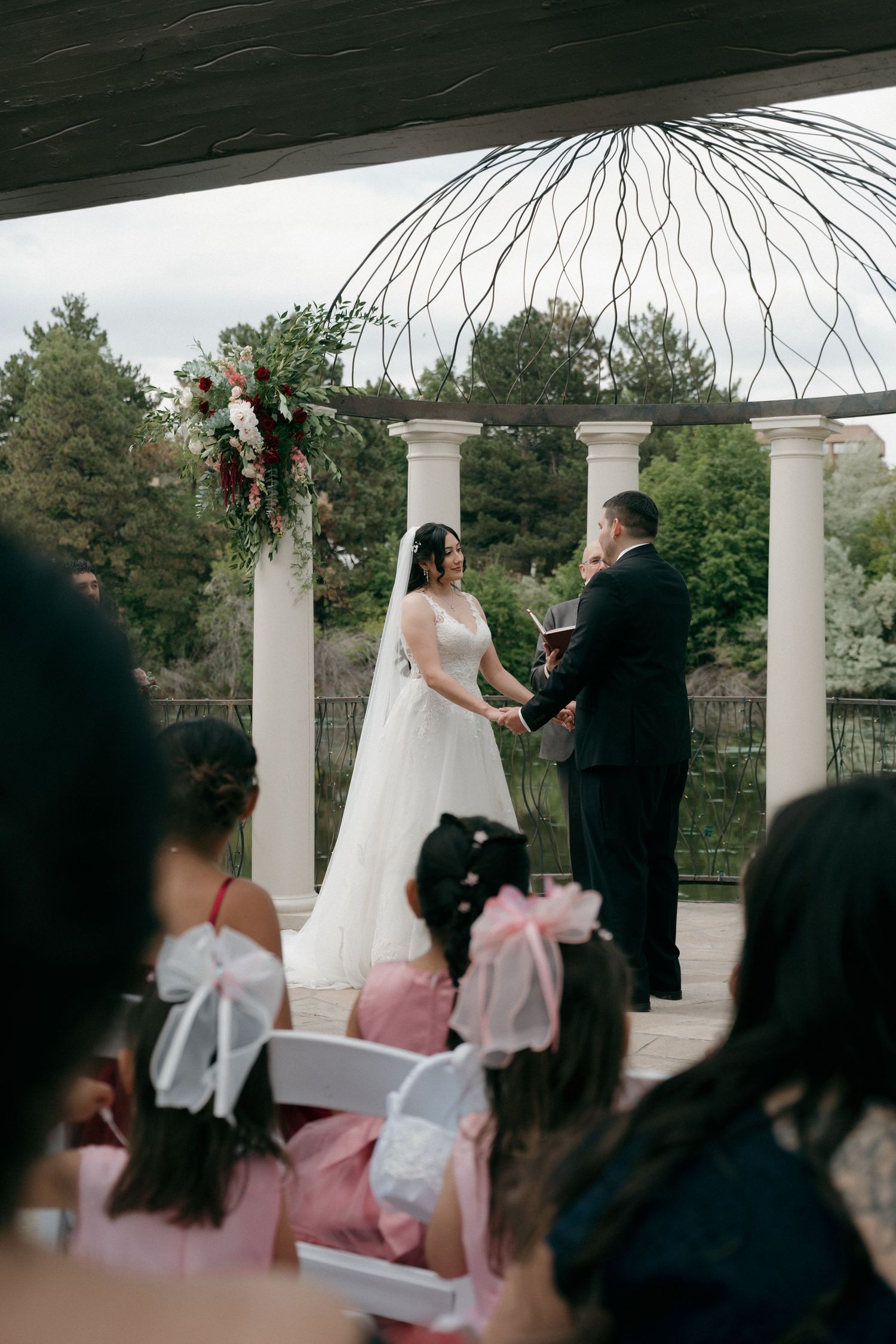 Bride and groom exchanging vows under floral arch at outdoor wedding ceremony with white columns.