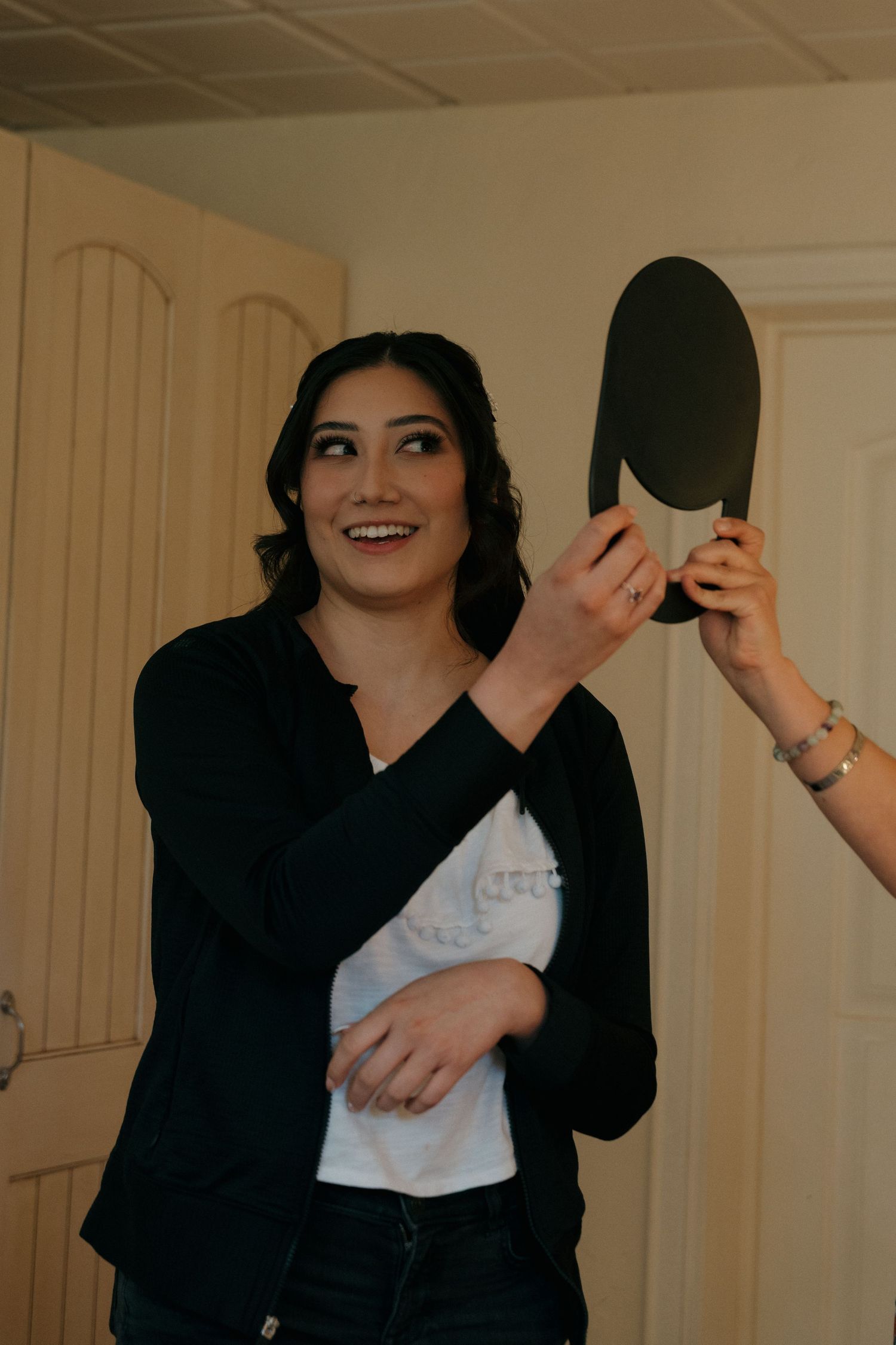 Smiling dark-haired woman in black cardigan looking into a handheld mirror during wedding preparations.