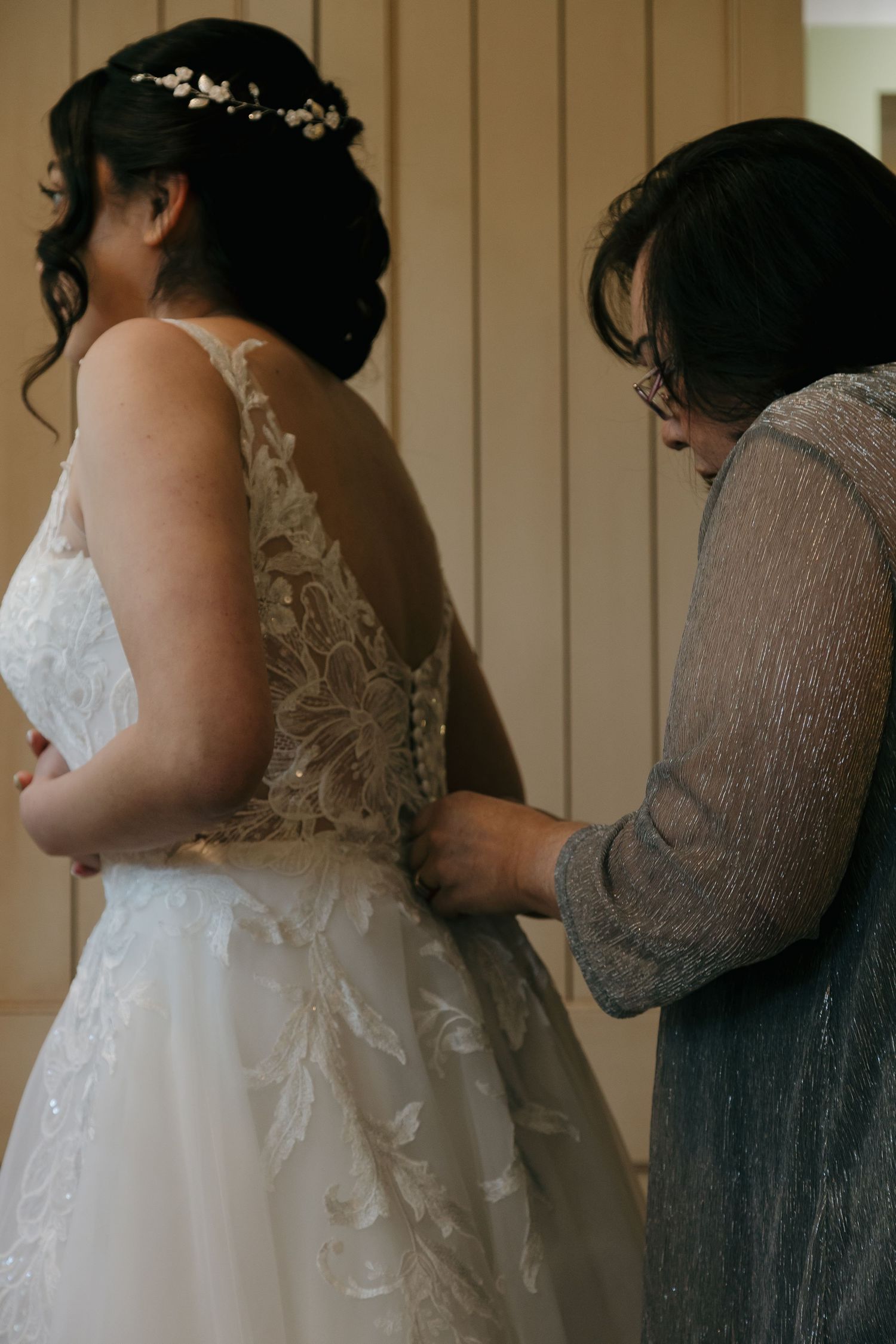 Mother helping bride fasten the back of a lace wedding gown during bridal preparations indoors.