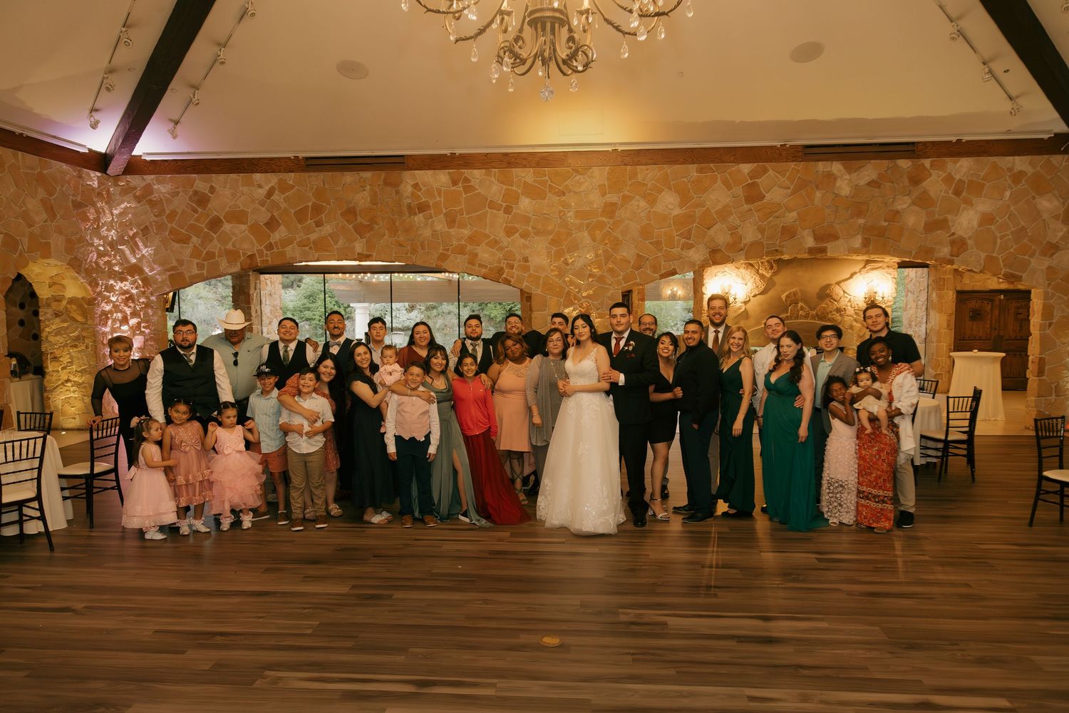 Large wedding family group photo with bride and groom in elegant rustic reception hall.