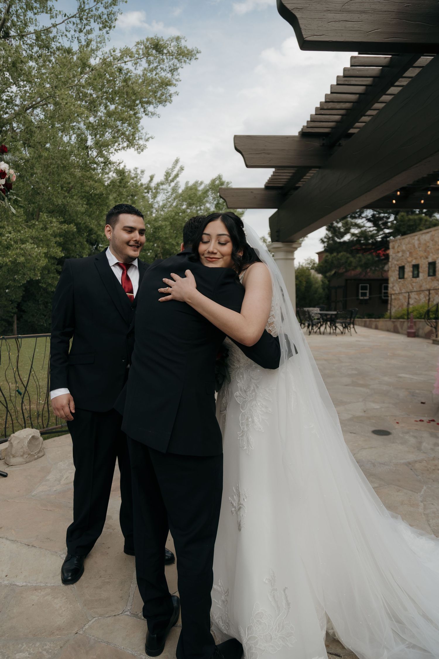 Bride in lace wedding gown embraces guest while groom in black suit with red tie stands nearby.