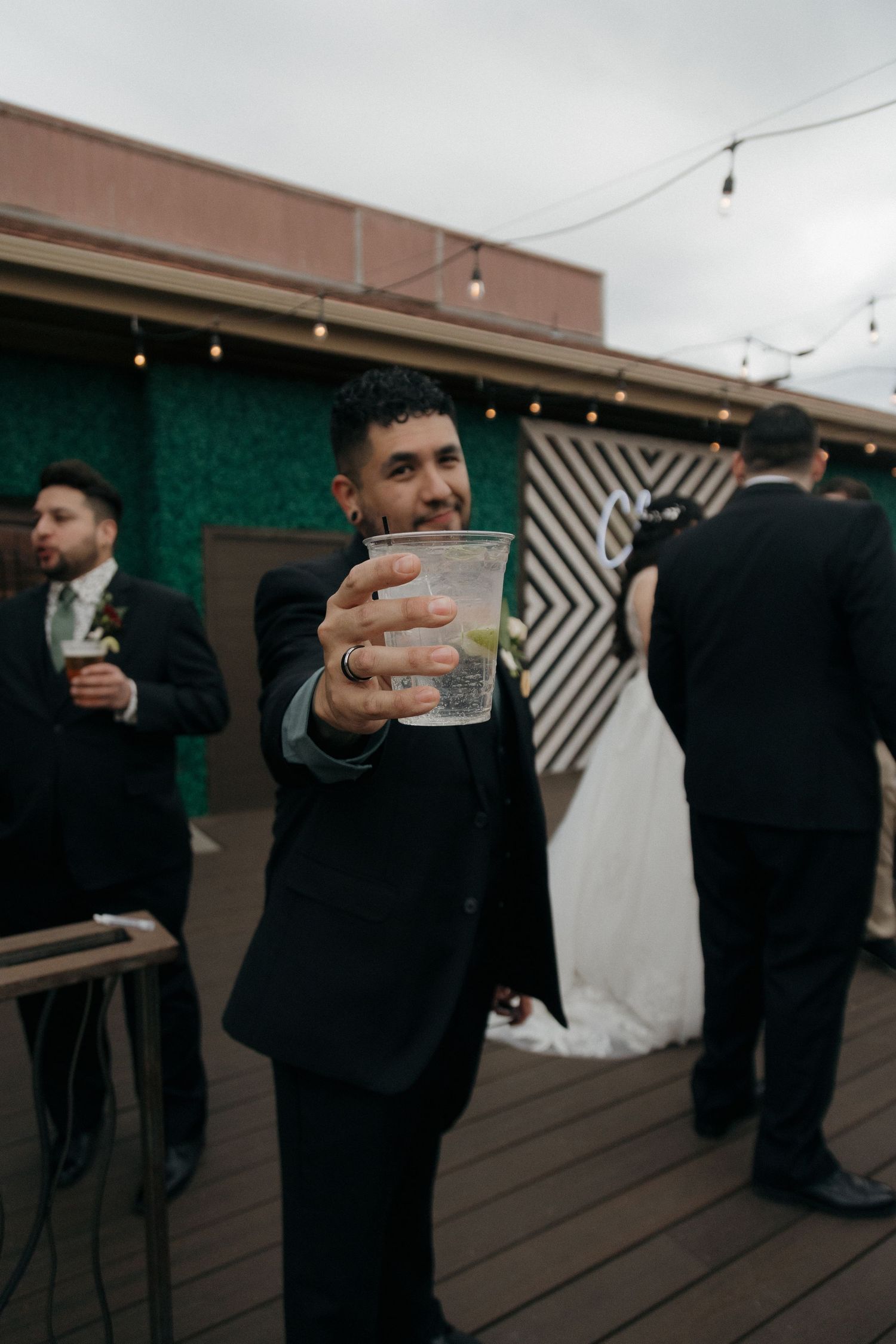 Smiling groomsman toasting with drink at wedding reception on outdoor deck with string lights.