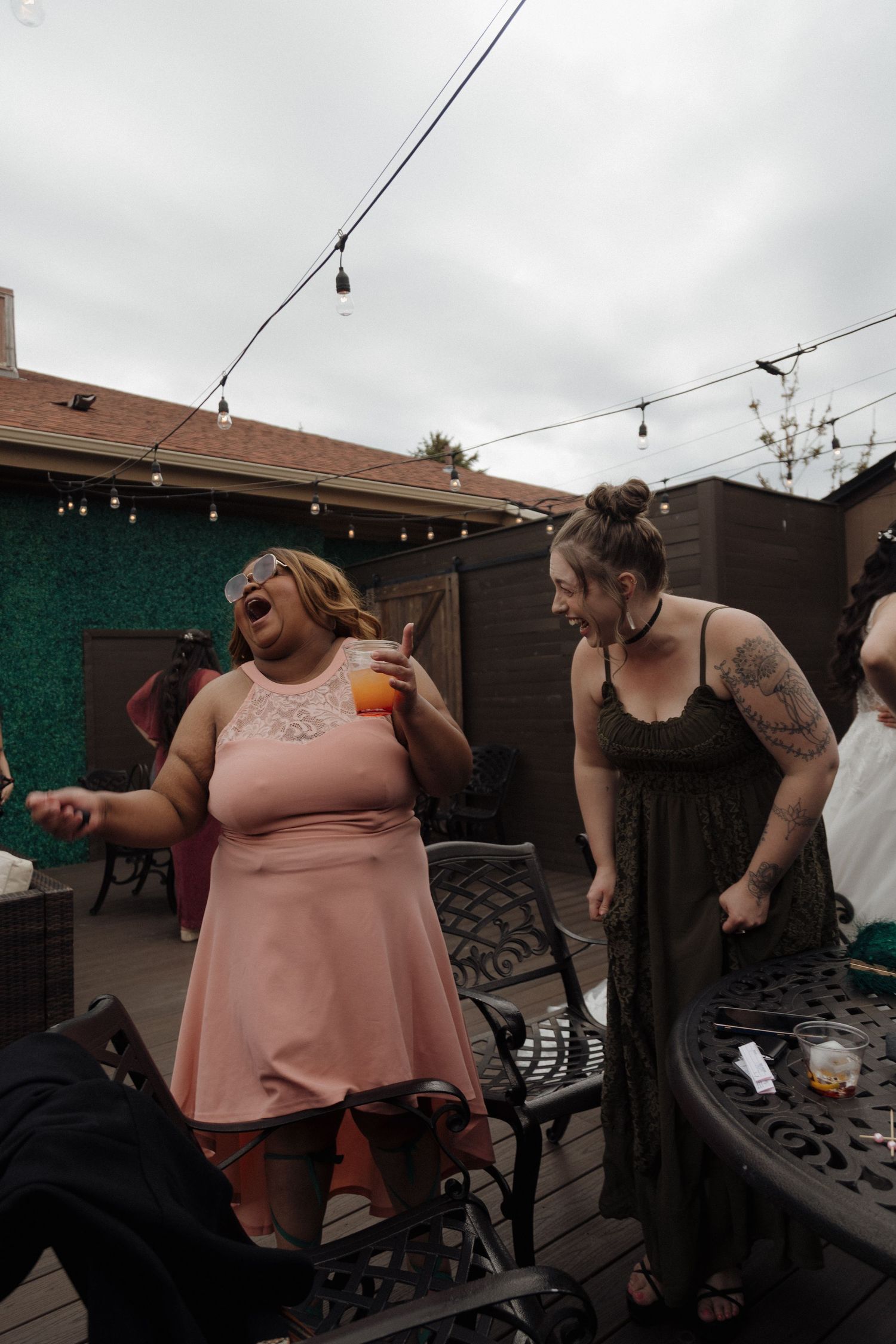 Two women laughing and dancing at outdoor wedding reception on deck with string lights overhead.