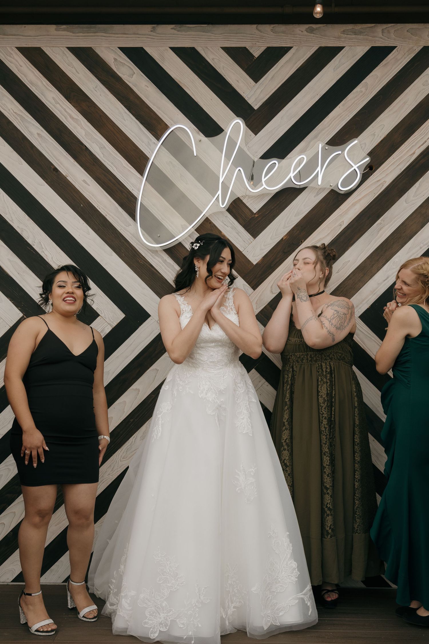 Bride laughing with bridesmaids in black and green dresses in front of a Cheers neon sign backdrop.