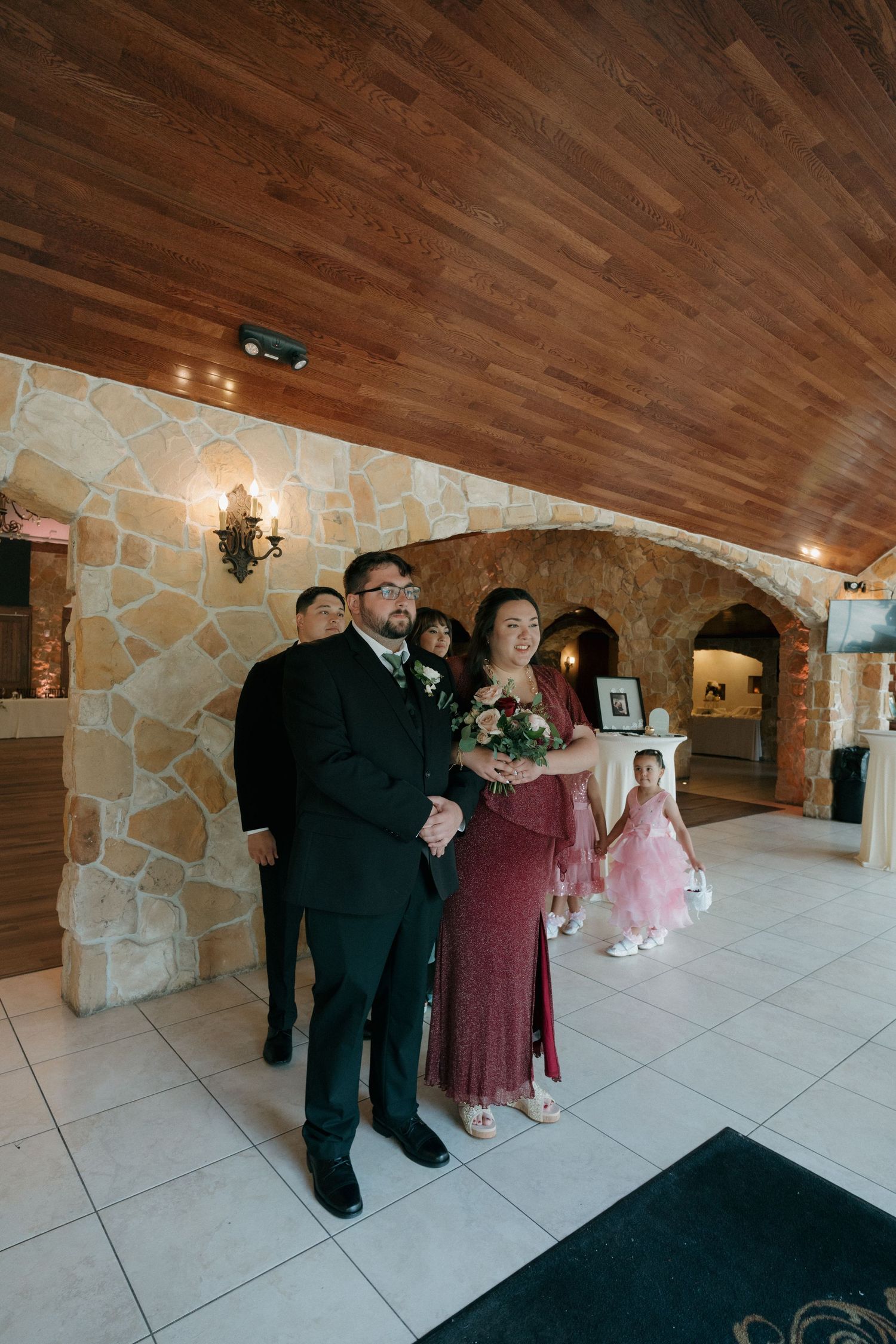 Bridesmaid in red dress walks arm-in-arm with groomsman through stone archway venue, flower girl follows behind.