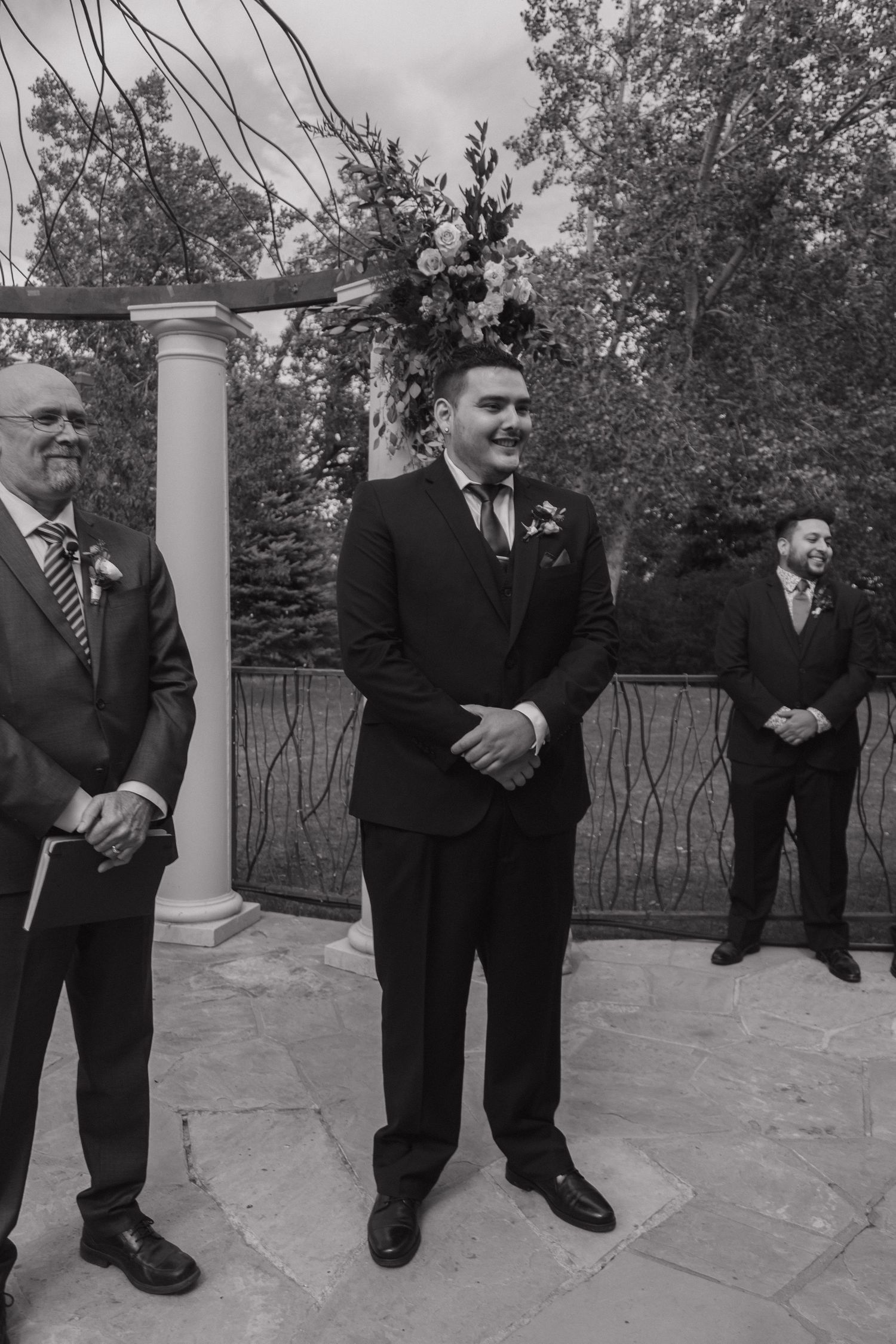 Smiling groom in black suit stands at the altar under a floral arch in a black and white photo.