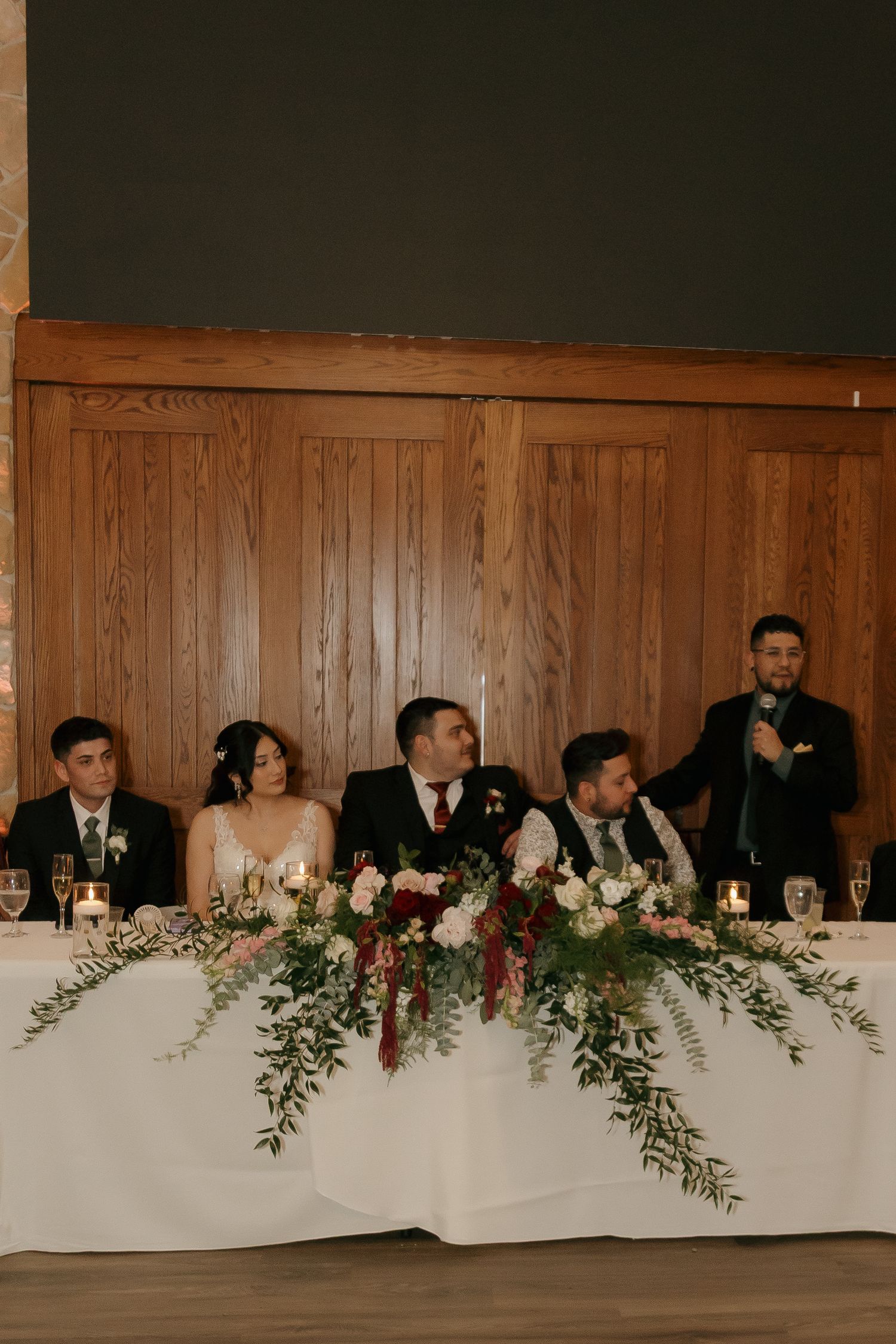 Wedding party seated at sweetheart table with lush greenery floral arrangements during reception speeches.