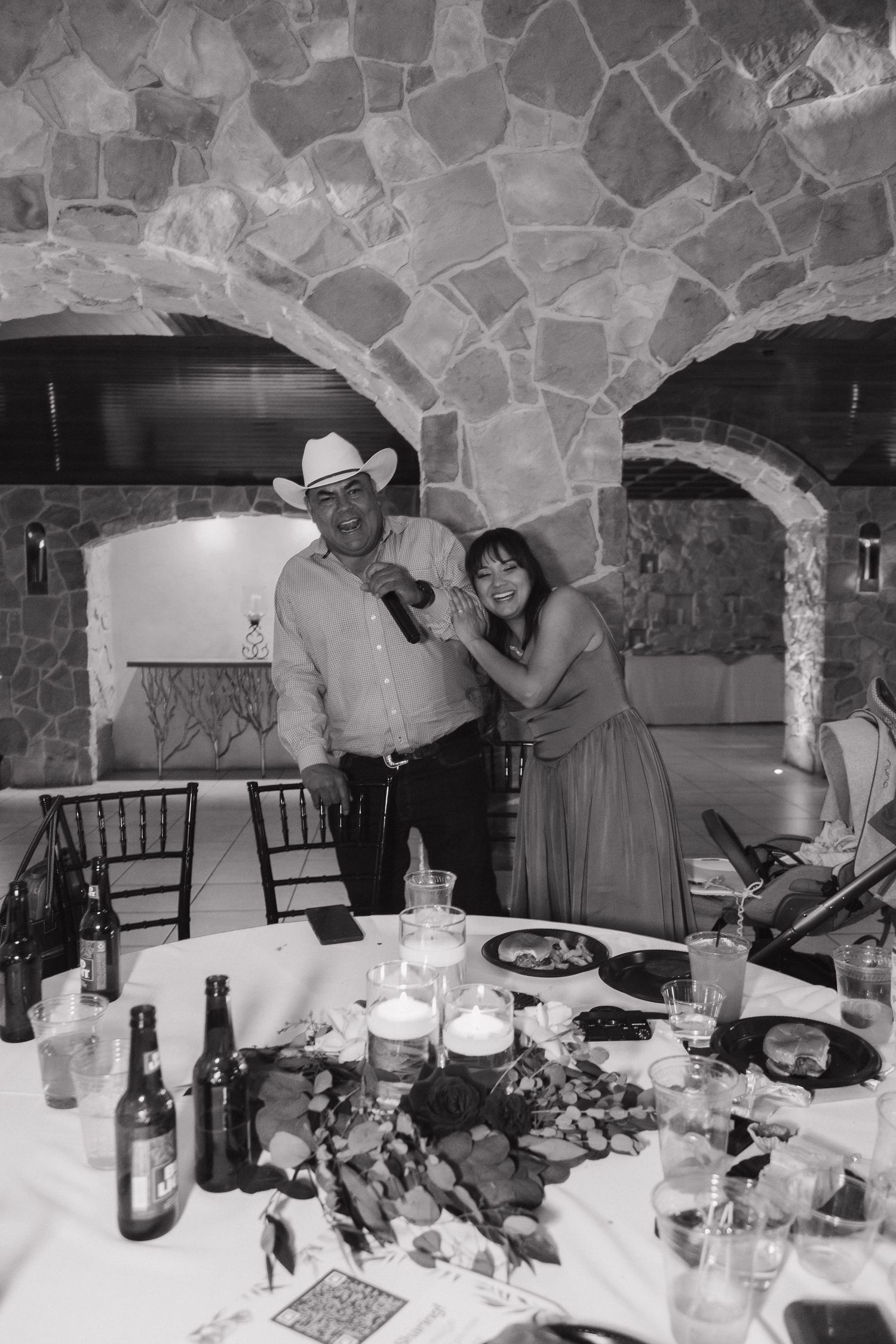 Couple smiling together at a wedding reception table with drinks, in a rustic stone-walled venue, black and white photo.