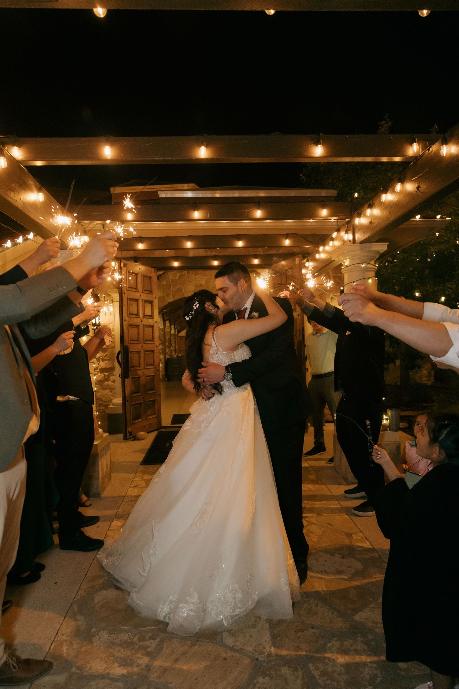 Newlyweds share a kiss under string lights surrounded by sparkler-holding guests at night.