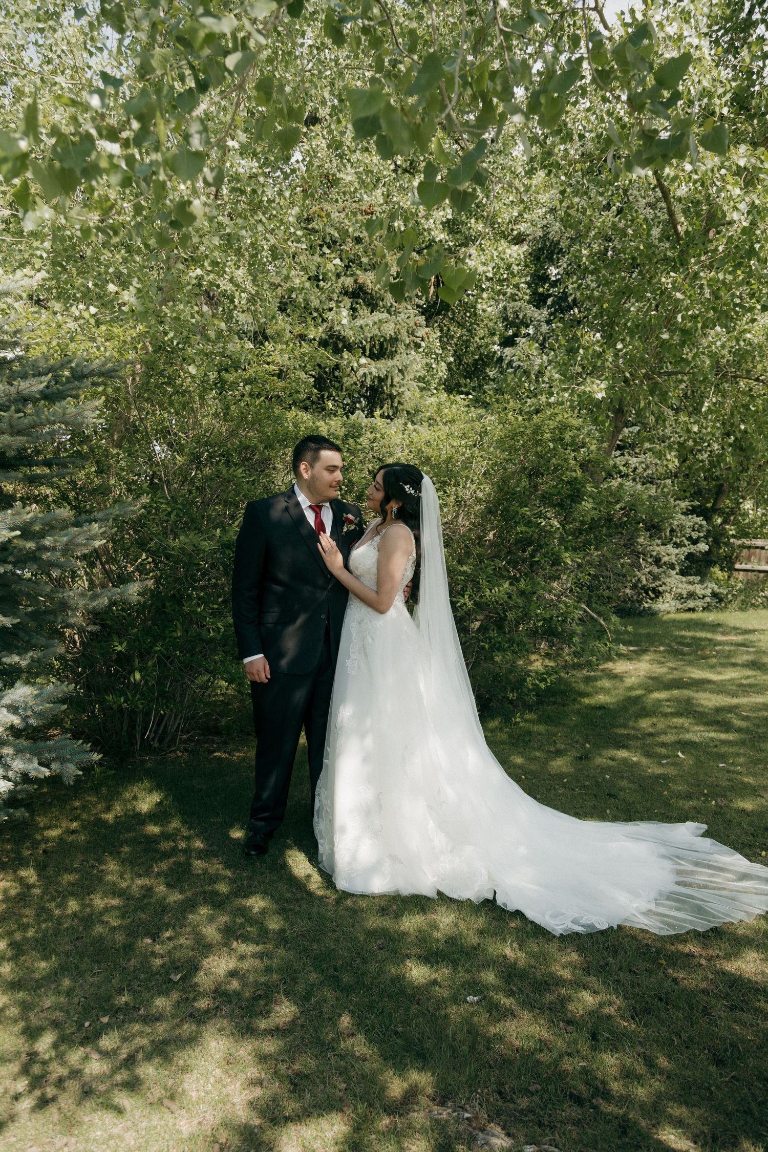 Bride and groom share a romantic moment surrounded by lush green trees in an outdoor garden setting.