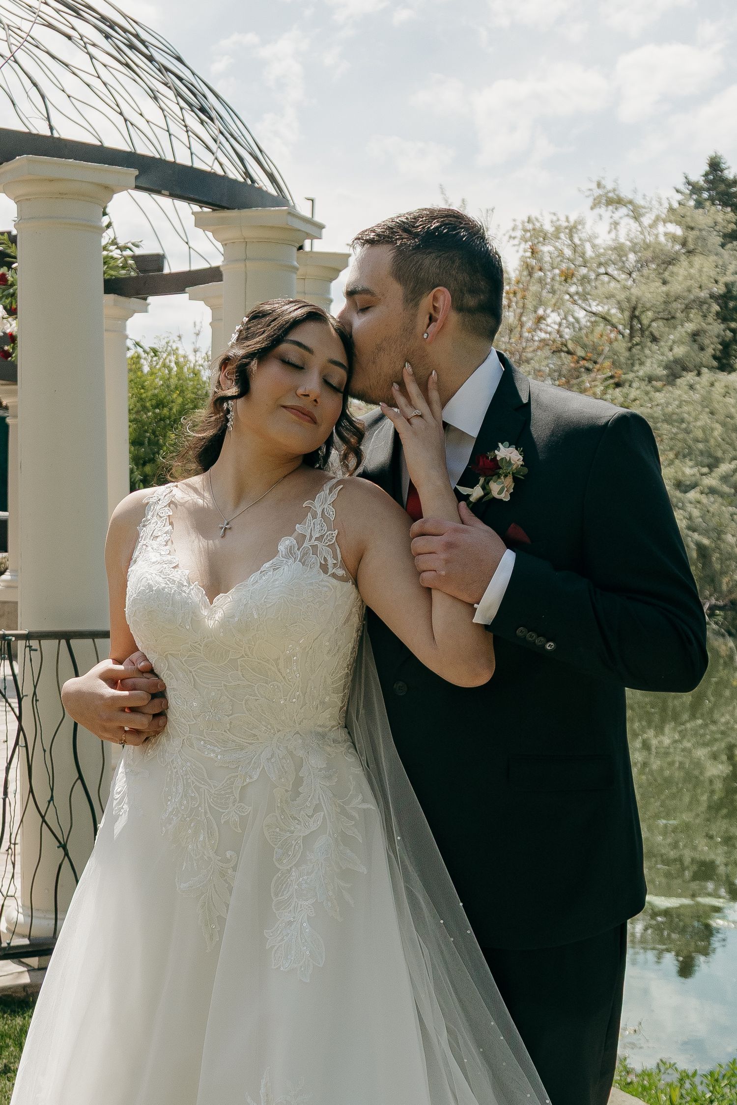 Groom in dark suit kisses bride on cheek near a gazebo and pond on their wedding day.