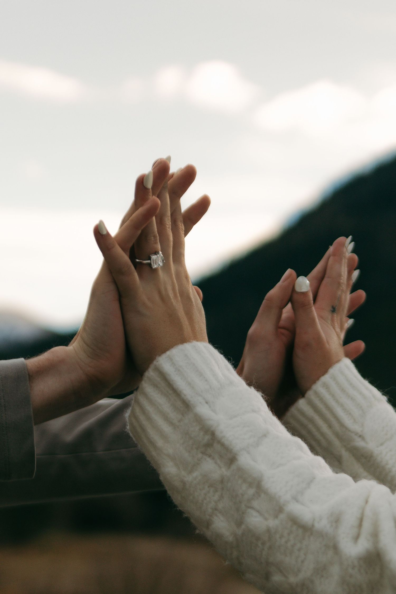 Two hands wearing white sweater sleeves reach up against a cloudy sky, with engagement rings visible on their fingers.