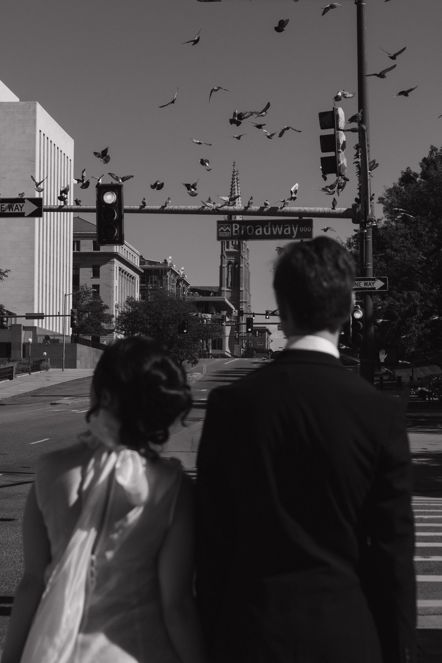 A black and white urban street scene with birds flying overhead as a couple waits at a crosswalk.