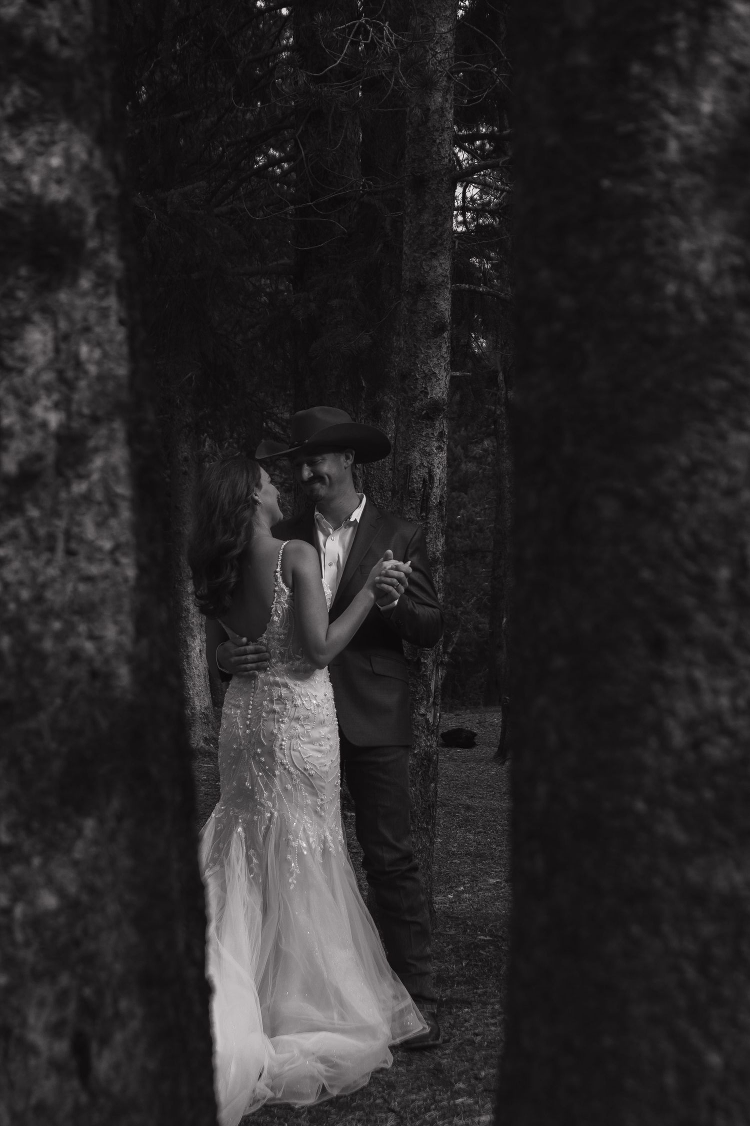 A romantic black and white wedding photo of a couple embracing between tall trees in a forest.