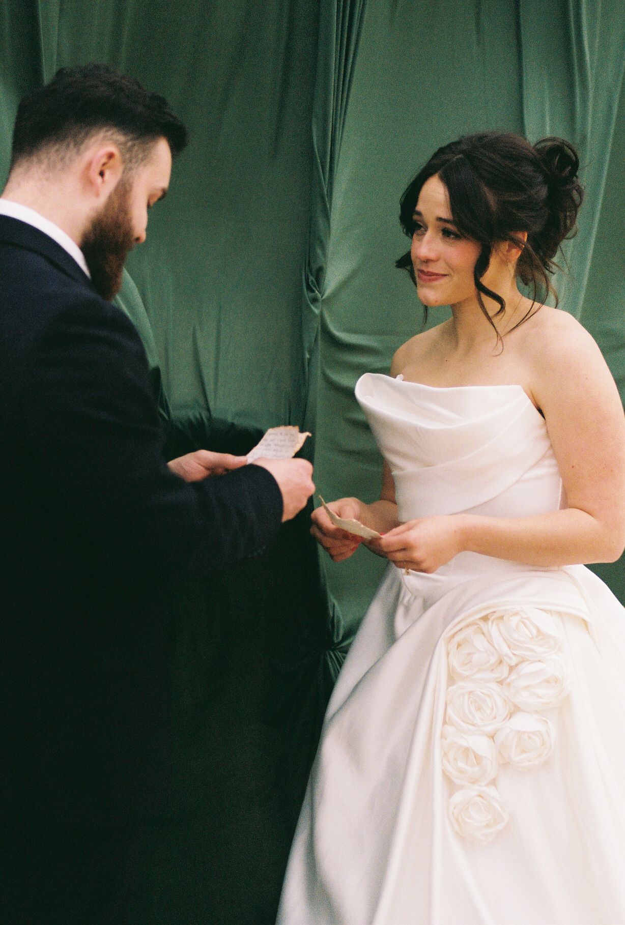 A couple exchanges rings during their wedding ceremony against a green curtain backdrop.