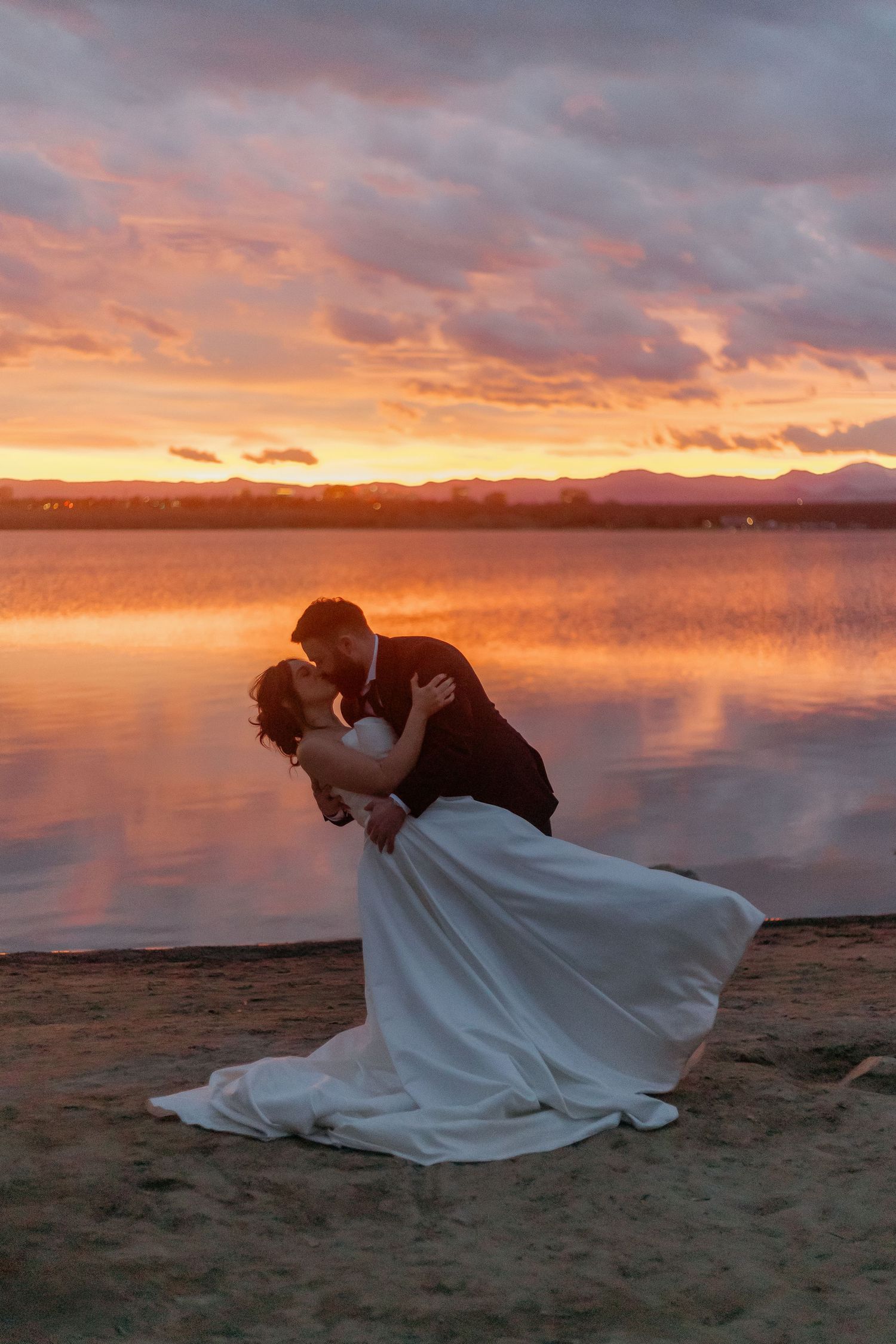A couple in wedding attire poses dramatically on a beach during a vibrant sunset.