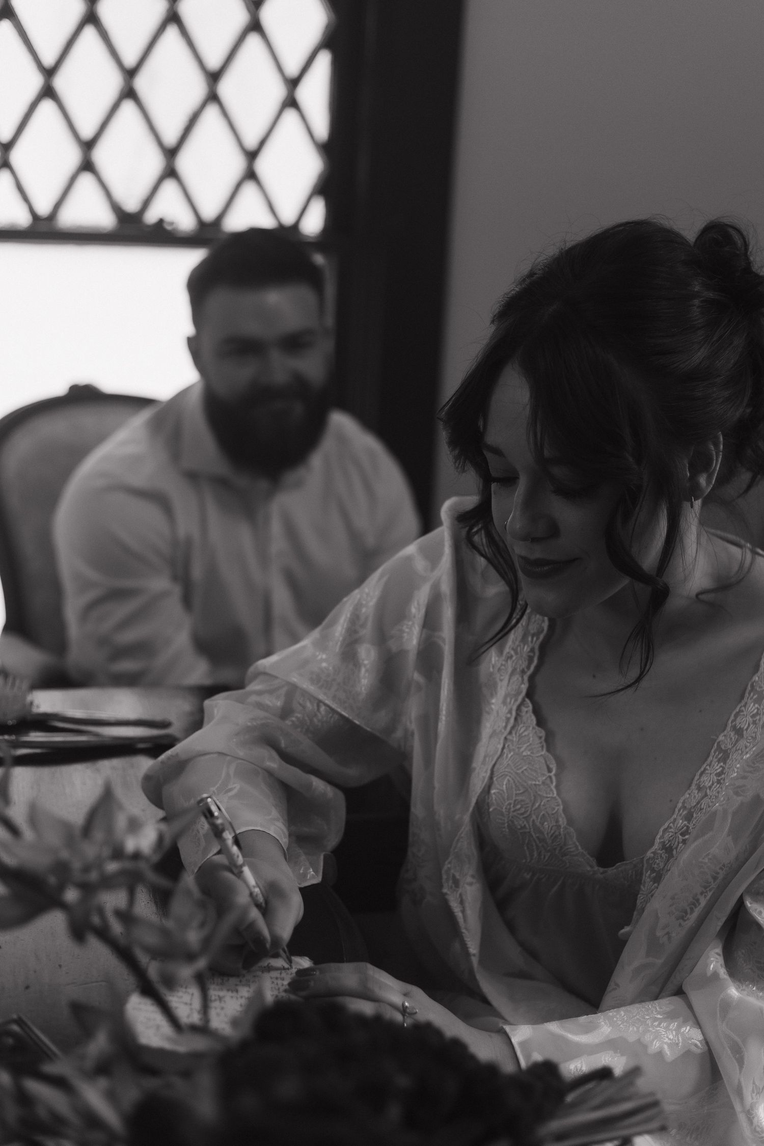 A couple signing documents at a wooden table near a window with diamond patterned glass.