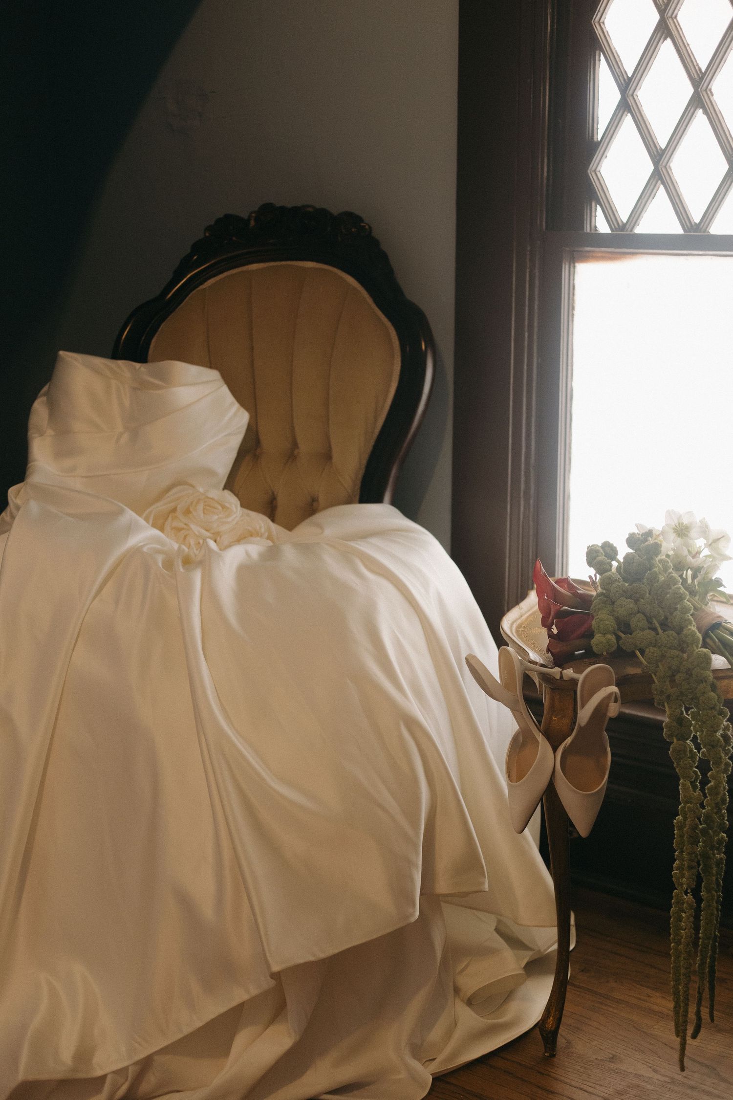 An elegant white wedding dress draped over a vintage chair near a window with diamond-patterned glass.