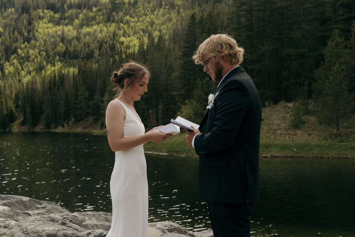 Bride and groom exchanging vows outdoors by a mountain lake surrounded by evergreen forest.