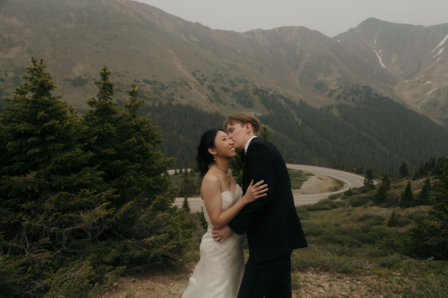 Bride and groom share a romantic kiss with stunning mountain scenery and evergreen trees backdrop.