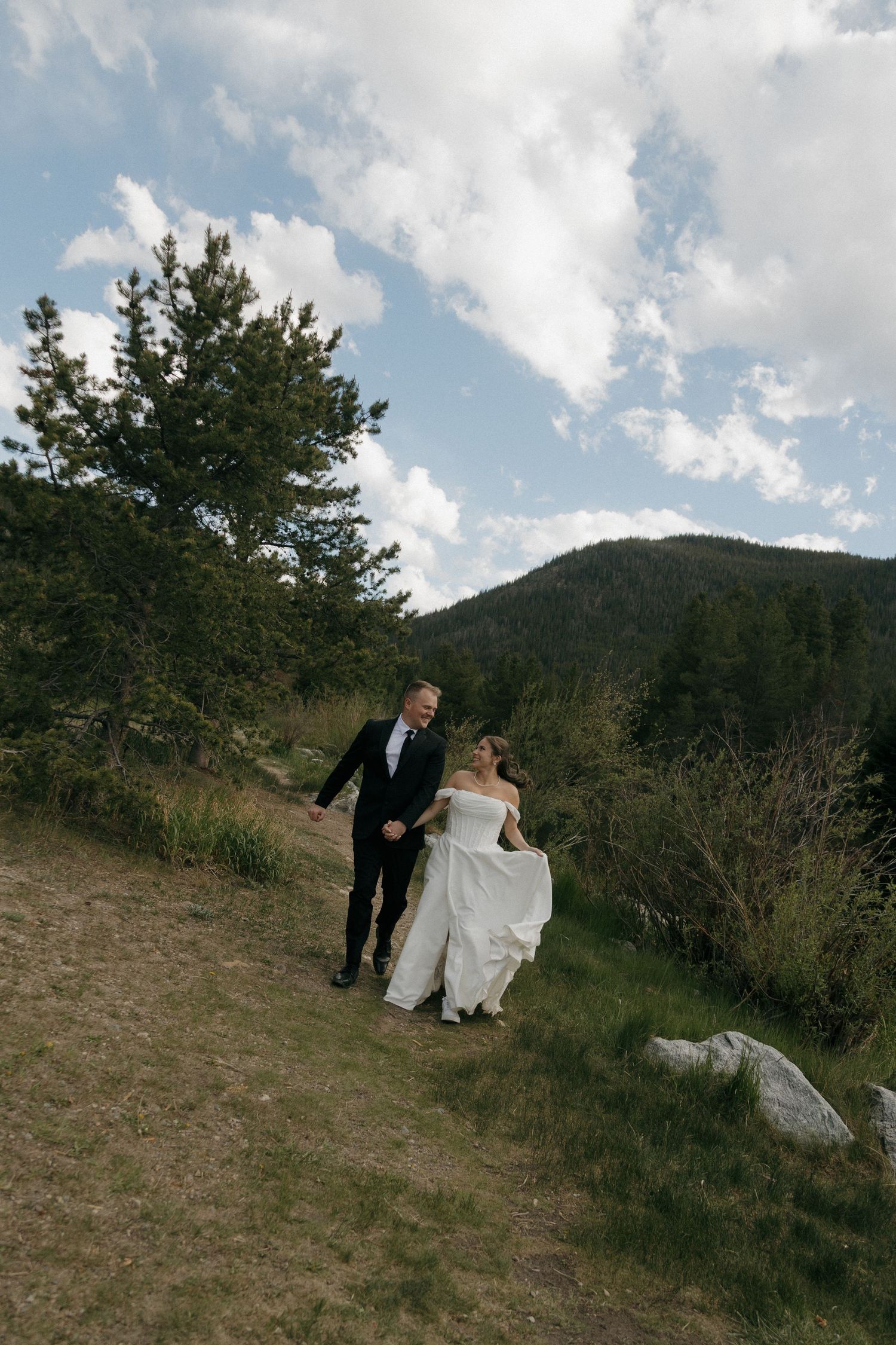 Bride in white off-shoulder gown and groom in black suit walk hand-in-hand on a mountain hillside.
